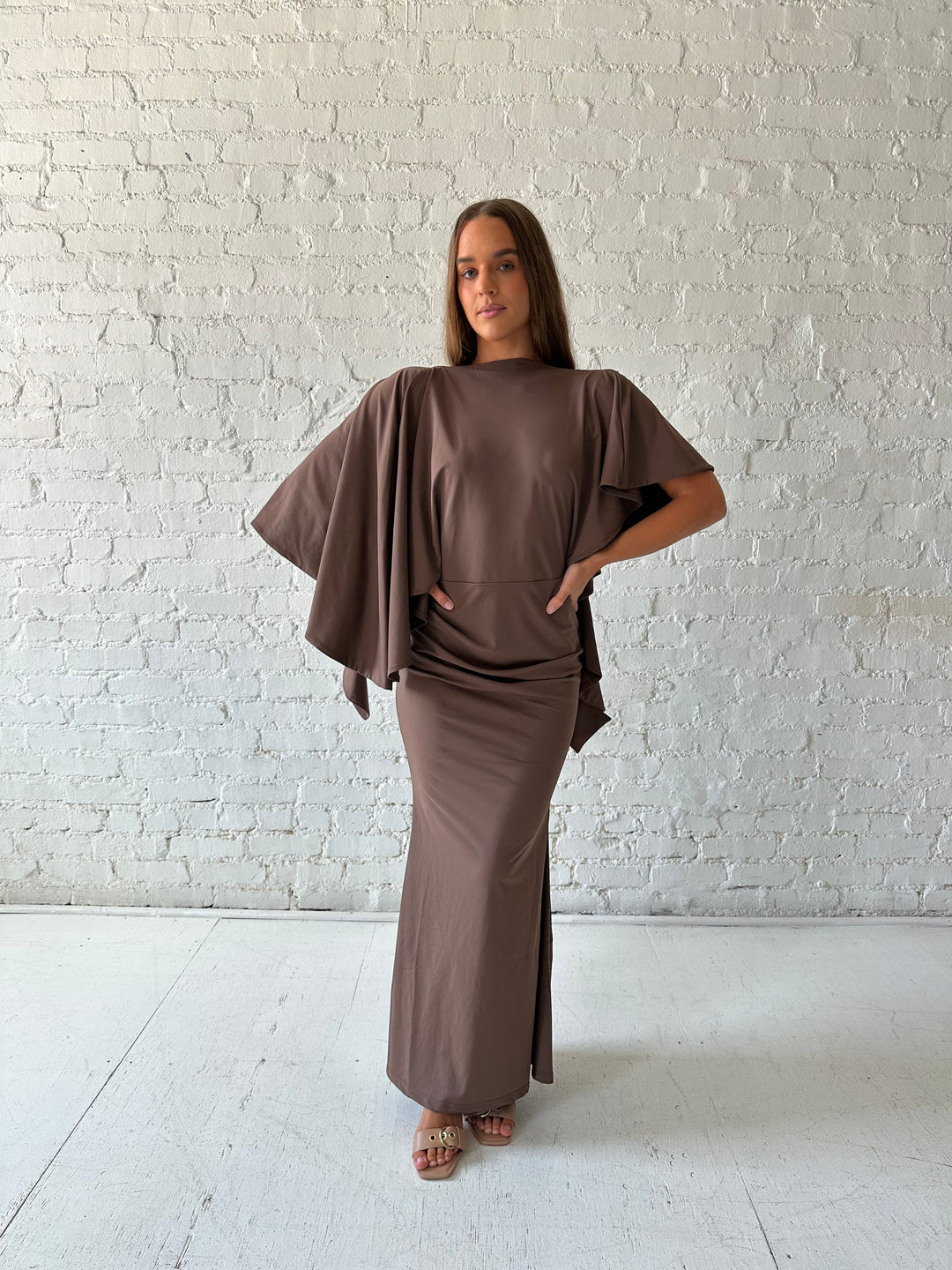 Woman wearing brown maxi dress with open back and flowy draped sleeves. 