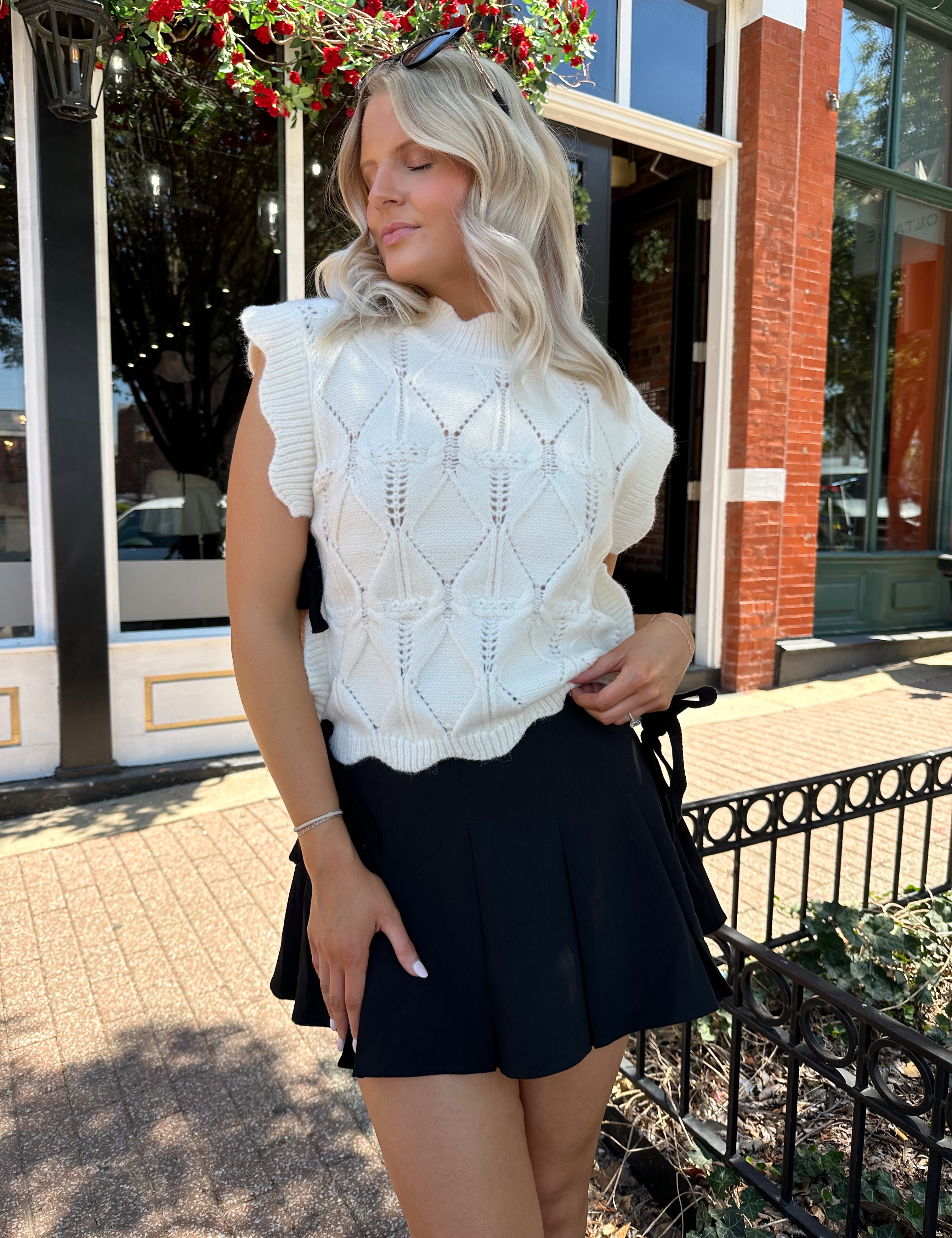 Woman wearing cream cable knit sleeveless sweater with black bow side detail. 