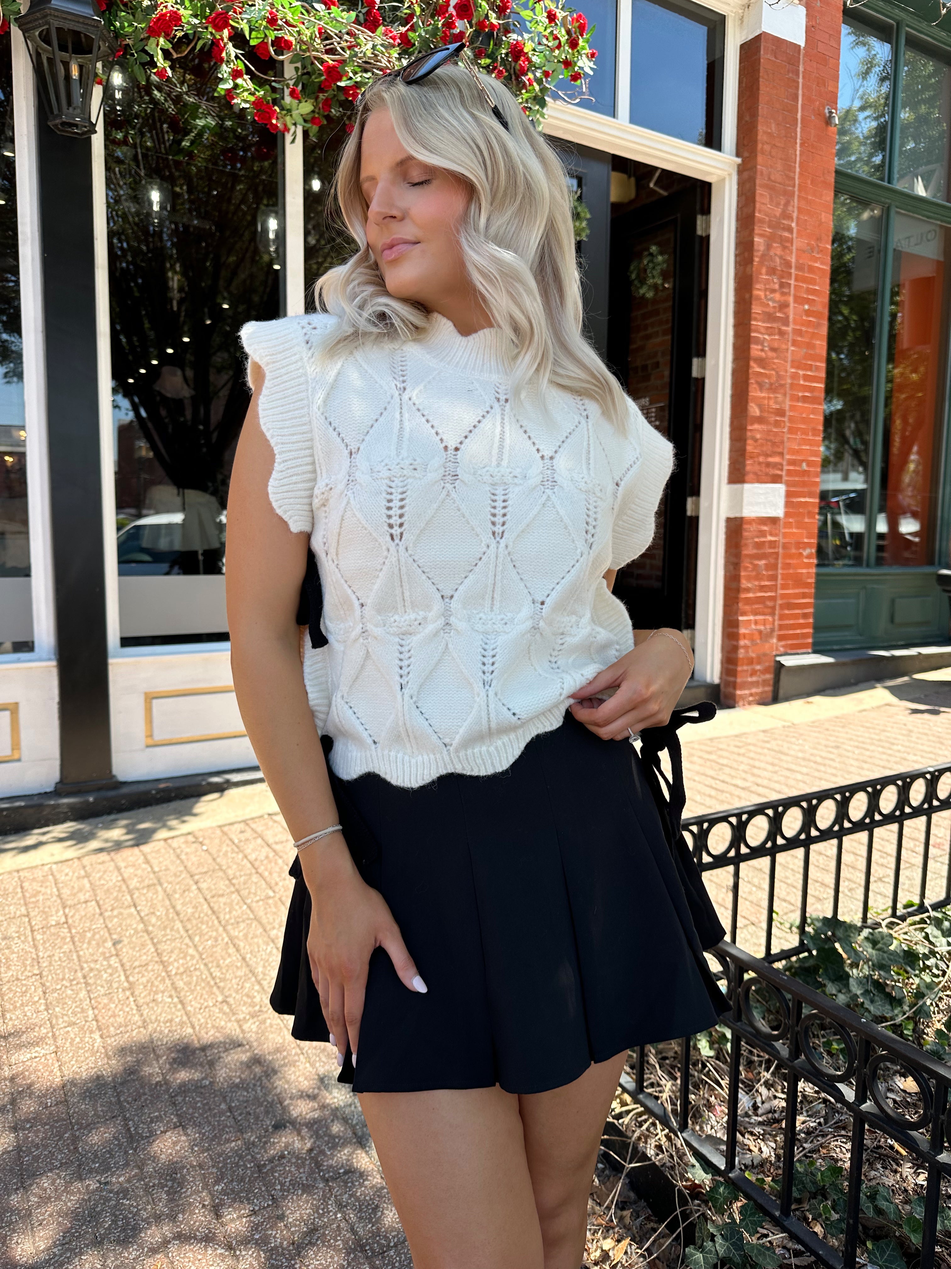 Woman wearing cream cable knit sleeveless sweater with black bow side detail. 