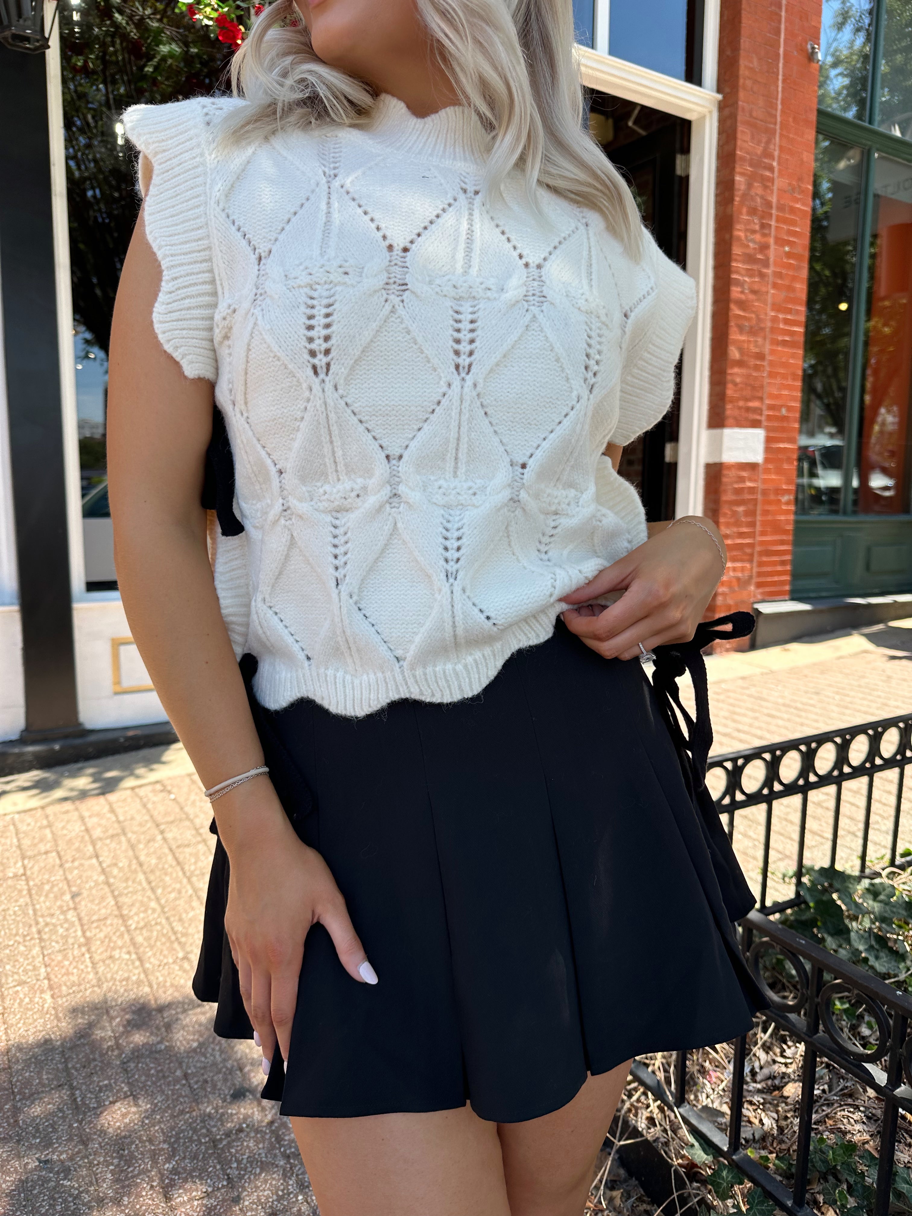 Woman wearing cream cable knit sleeveless sweater with black bow side detail. 