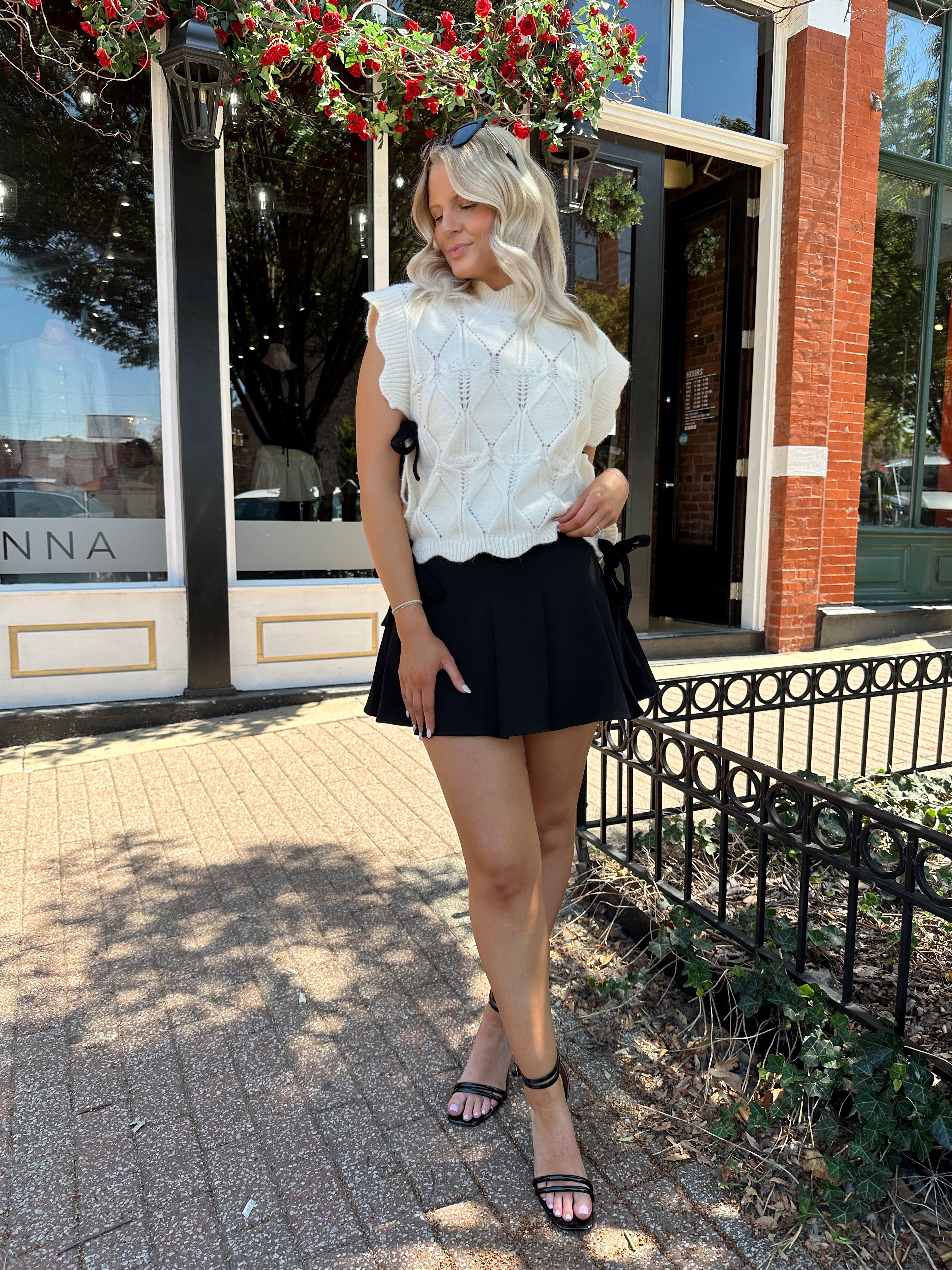 Woman wearing cream cable knit sleeveless sweater with black bow side detail. 