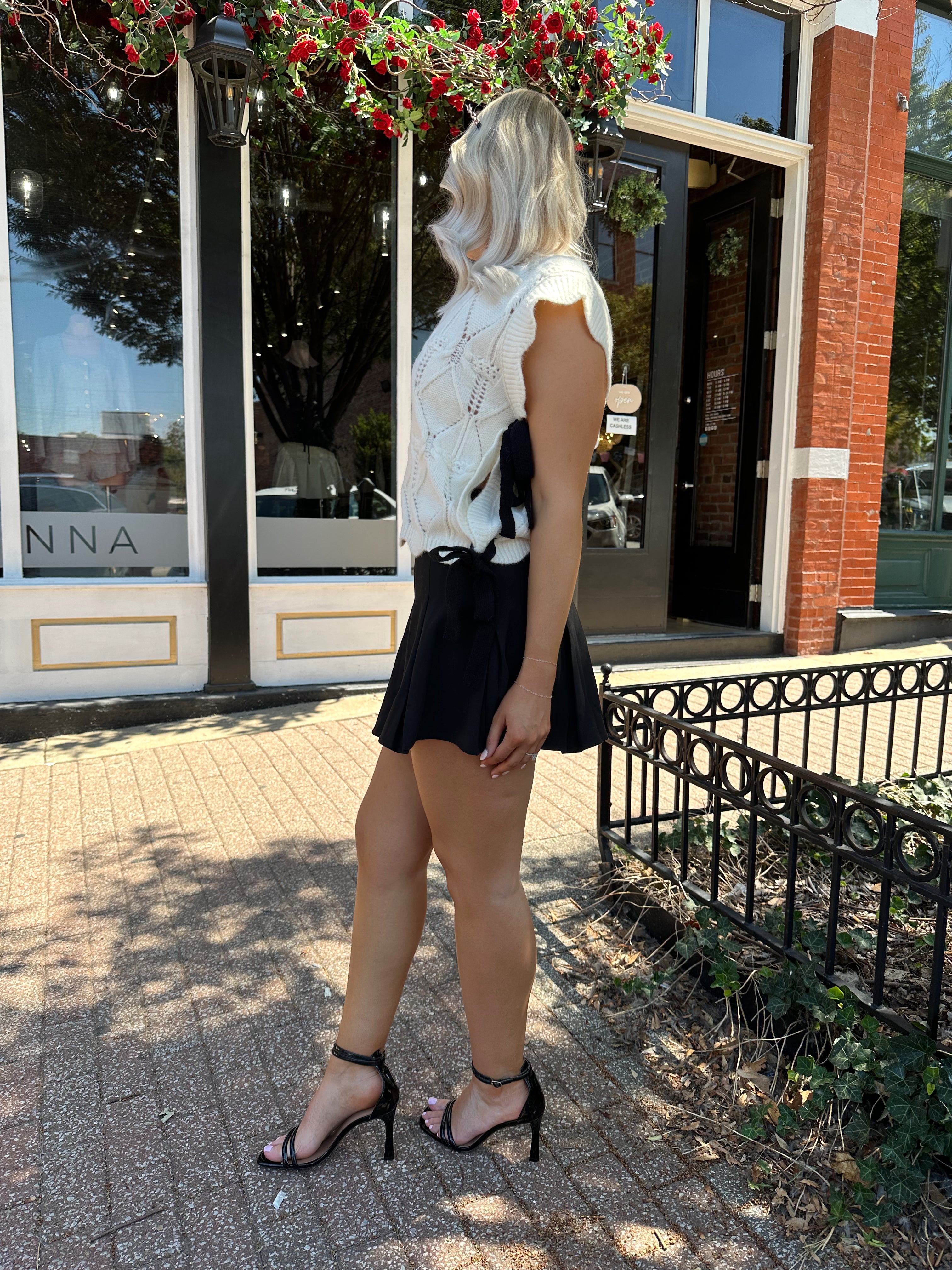Woman wearing cream cable knit sleeveless sweater with black bow side detail. 