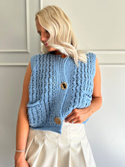 Woman wearing a light blues chunky sleeveless sweater with gold button closure.