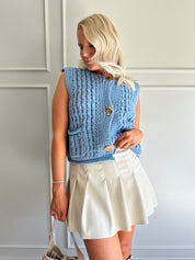 Woman wearing a light blues chunky sleeveless sweater with gold button closure.