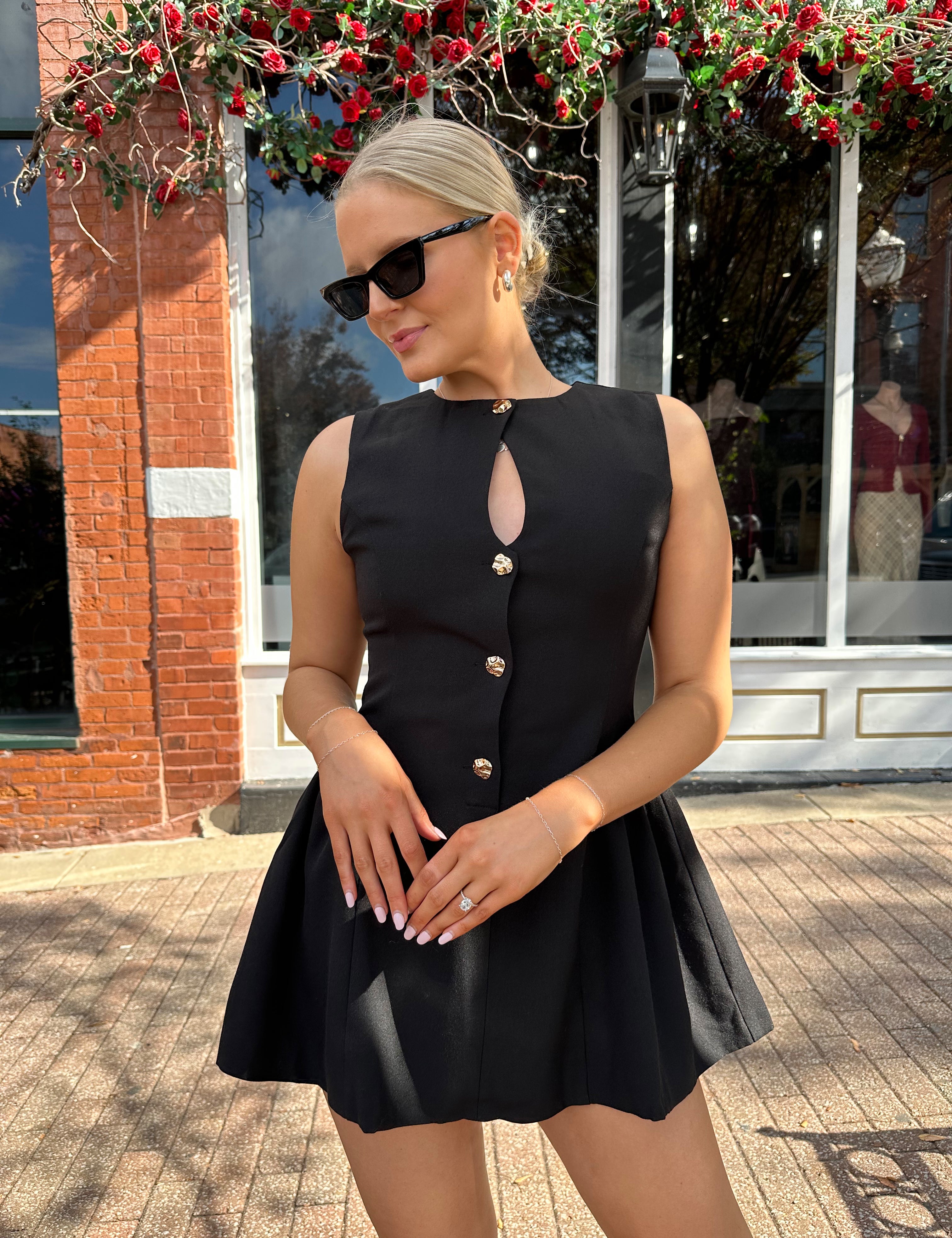 Woman wearing fit to flare black mini dress with keyhole cutout and gold buttons.