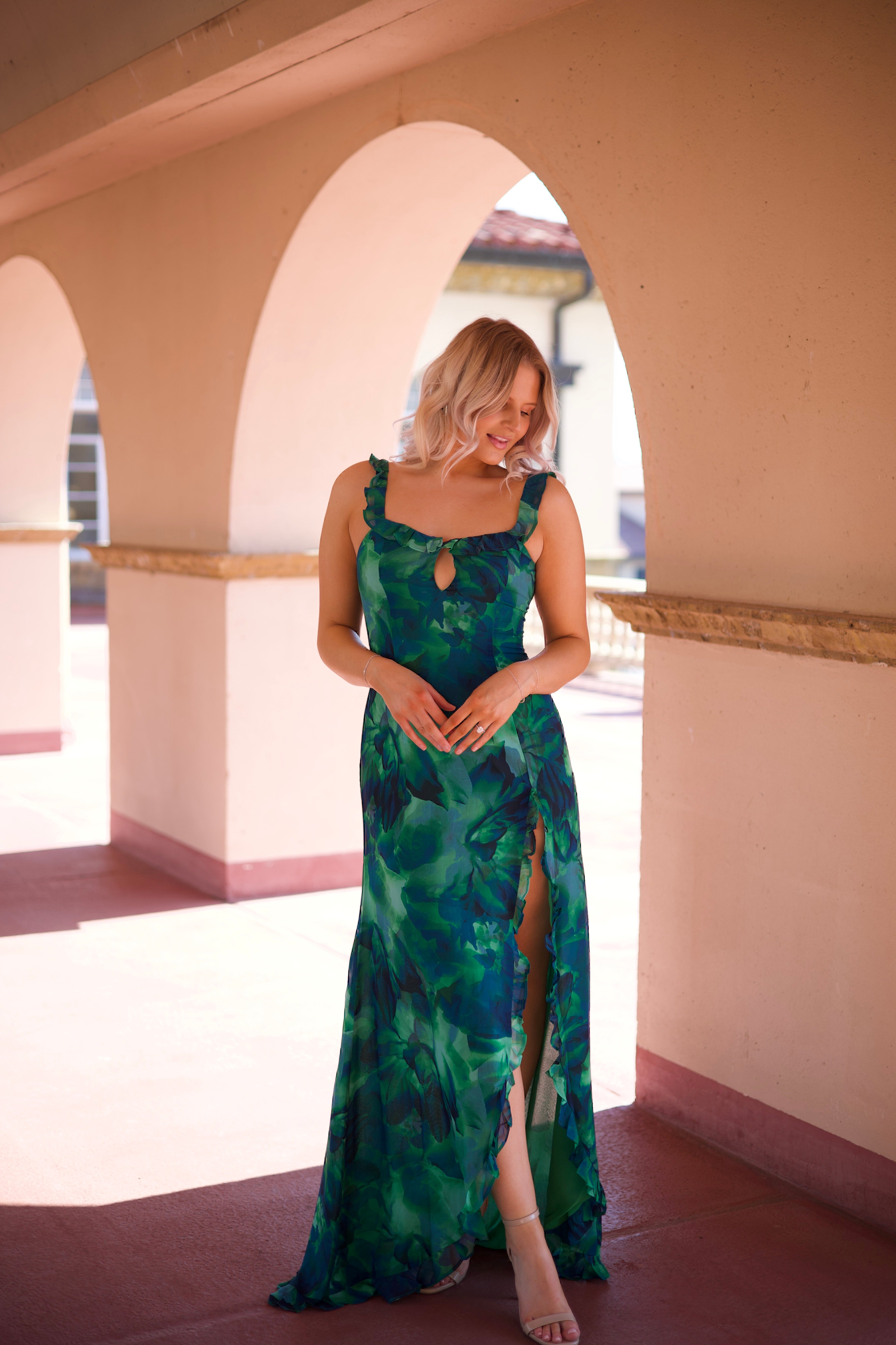 Calypso Tropical Maxi Dress with a vibrant green print, keyhole detail, and a side slit, walking under an arched terrace.