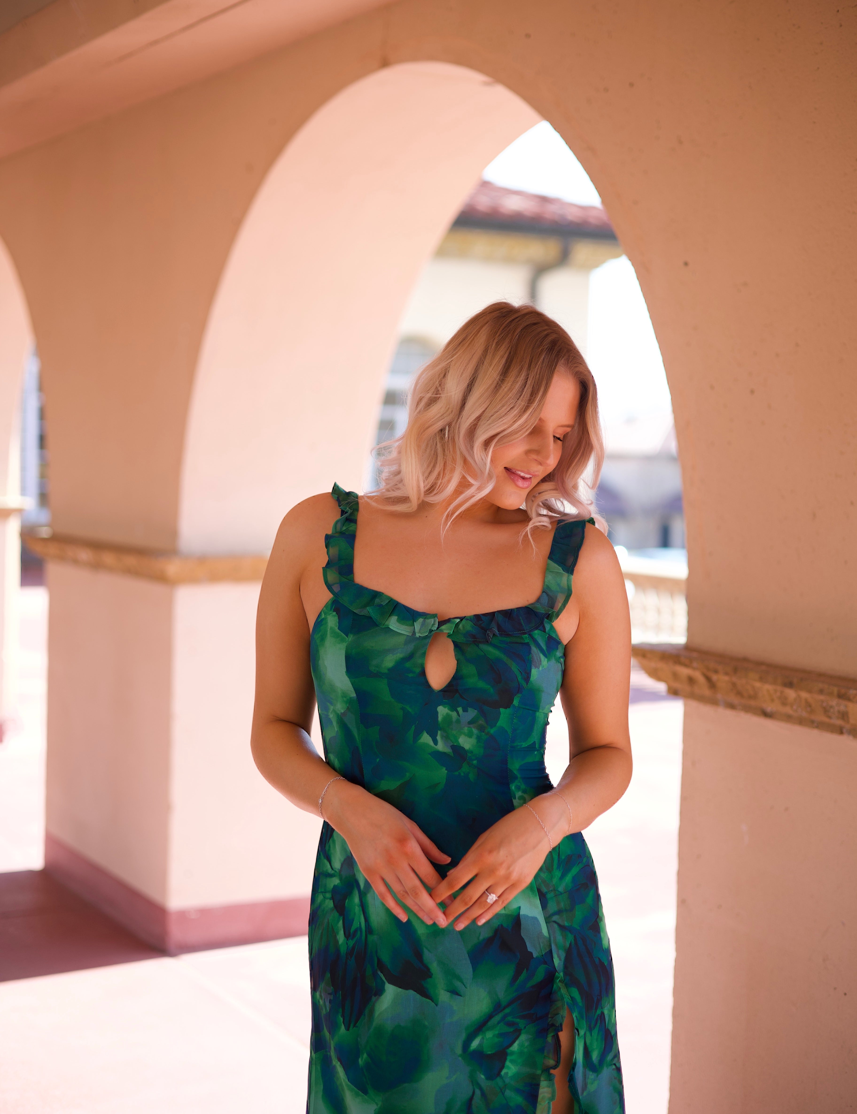 Calypso Tropical Maxi Dress with a vibrant green print, keyhole detail, and a side slit, walking under an arched terrace.