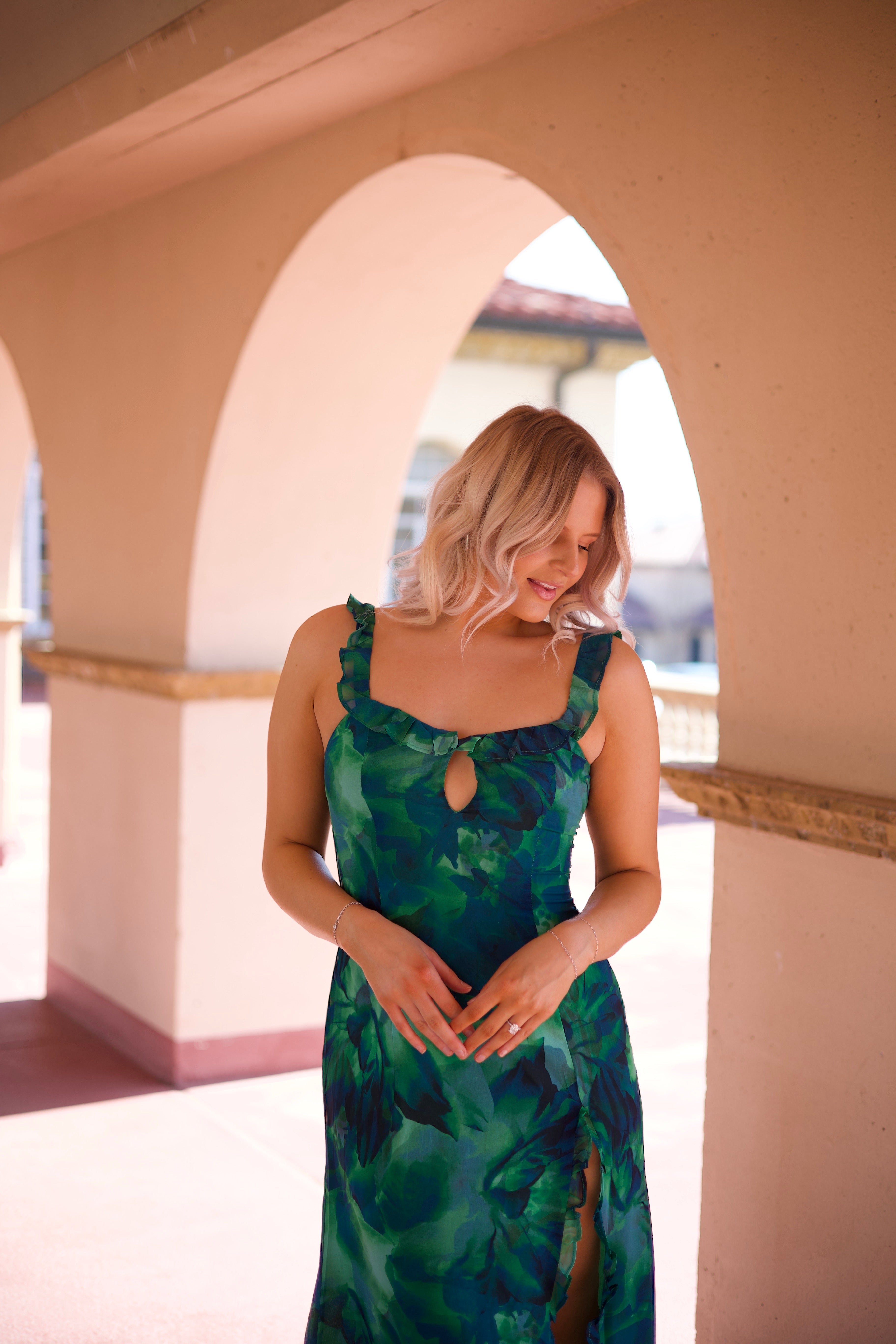 Calypso Tropical Maxi Dress with a vibrant green print, keyhole detail, and a side slit, walking under an arched terrace.