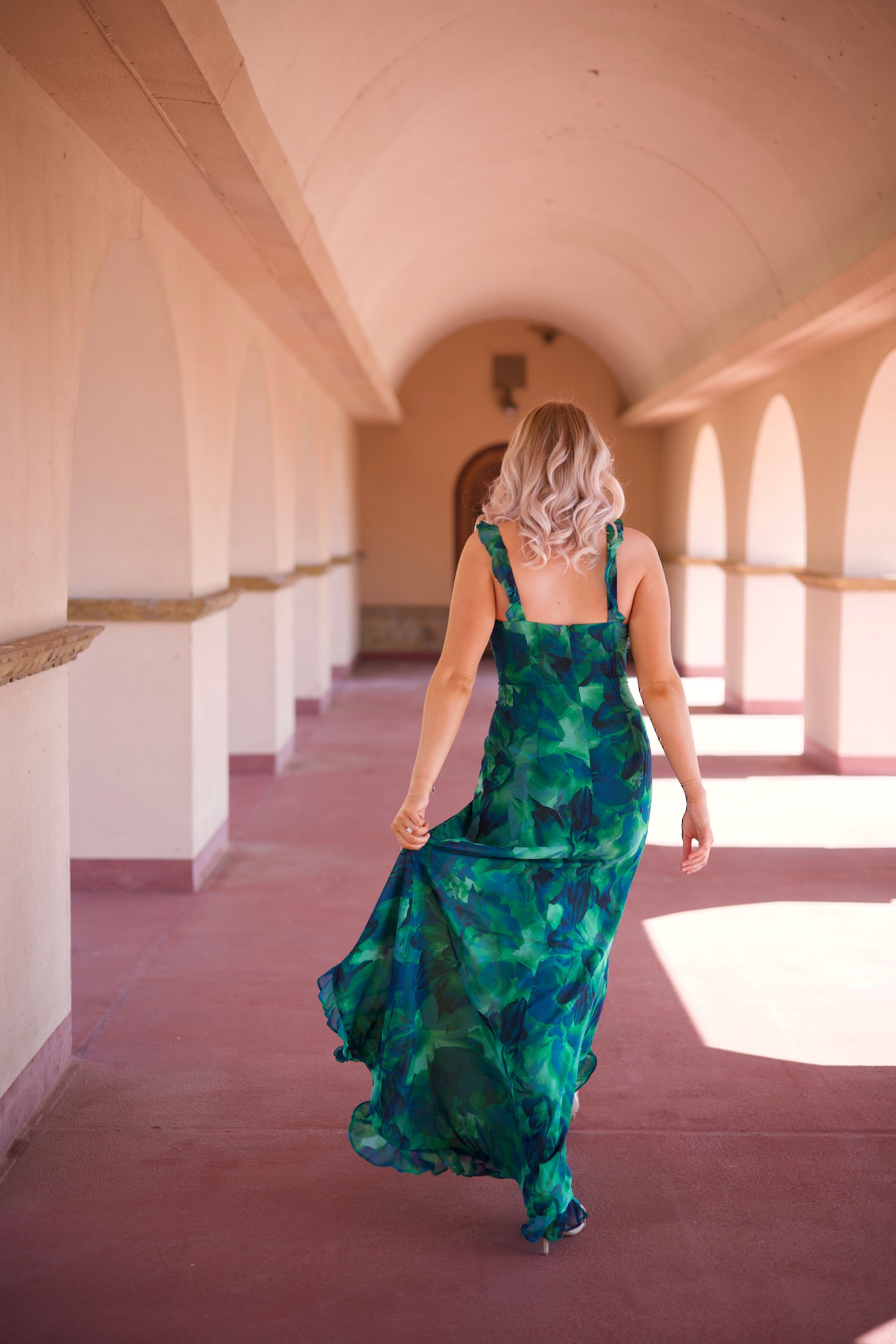 Calypso Tropical Maxi Dress with a vibrant green print, keyhole detail, and a side slit, walking under an arched terrace.