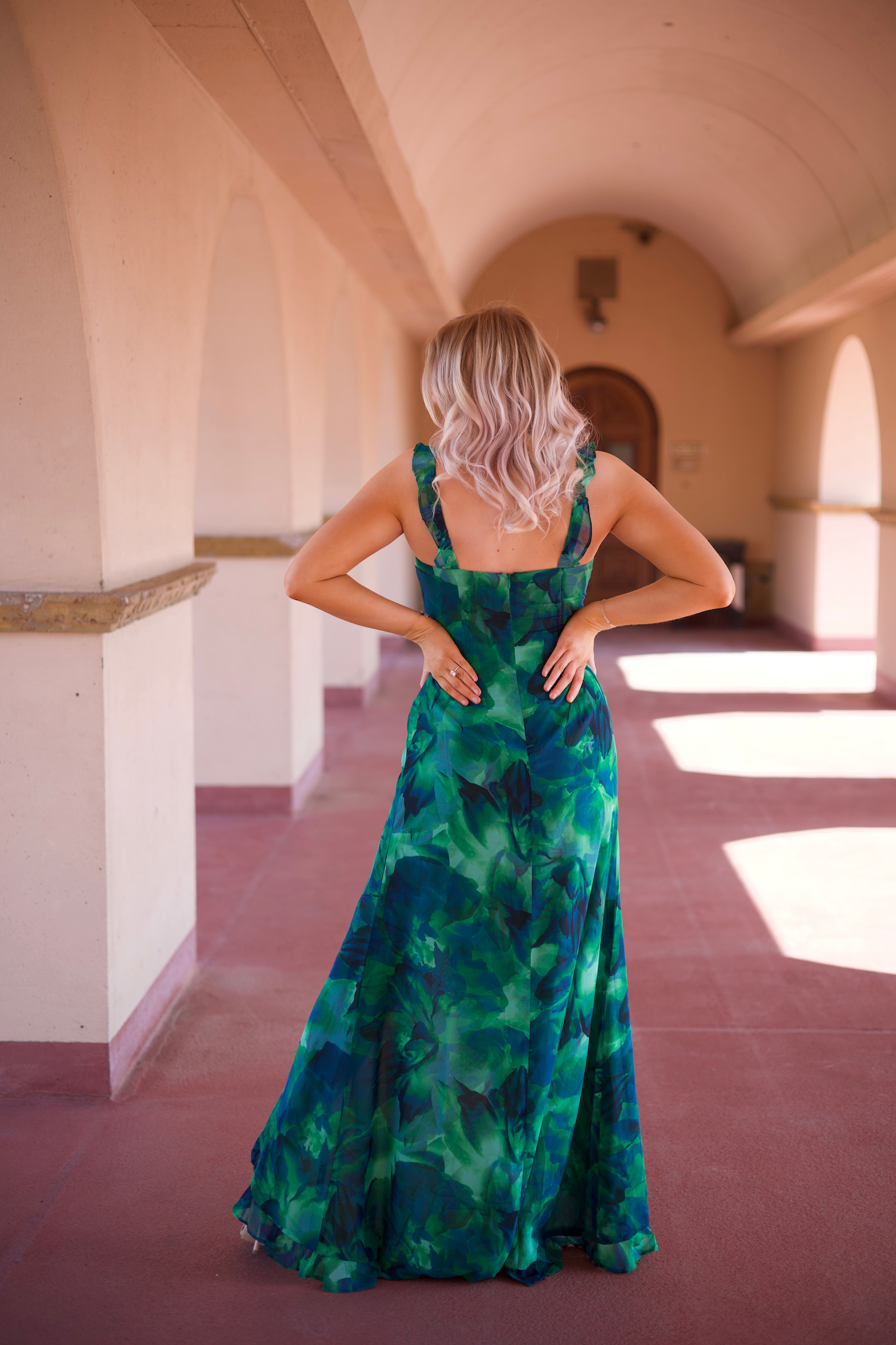 Calypso Tropical Maxi Dress with a vibrant green print, keyhole detail, and a side slit, walking under an arched terrace.