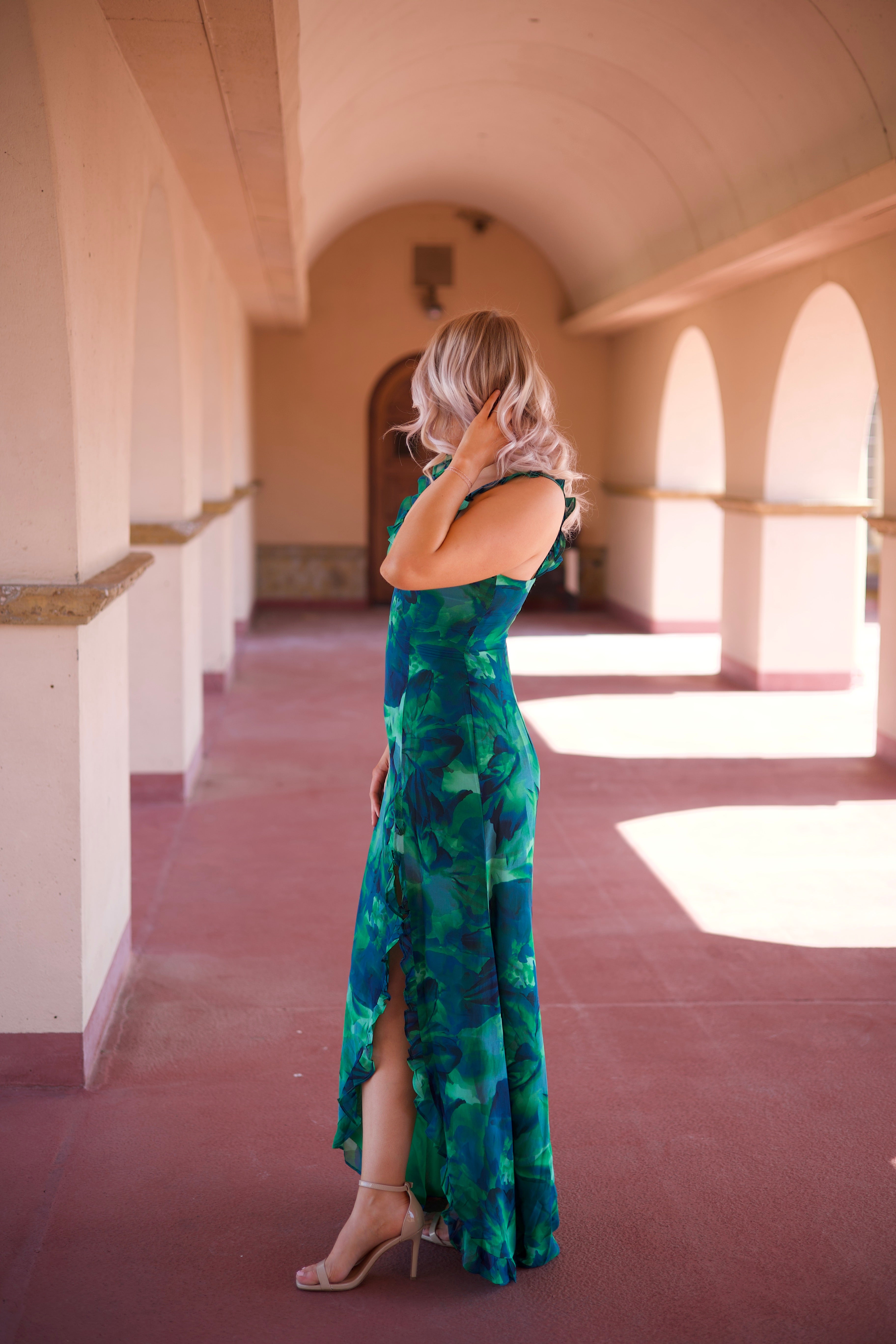 Calypso Tropical Maxi Dress with a vibrant green print, keyhole detail, and a side slit, walking under an arched terrace.