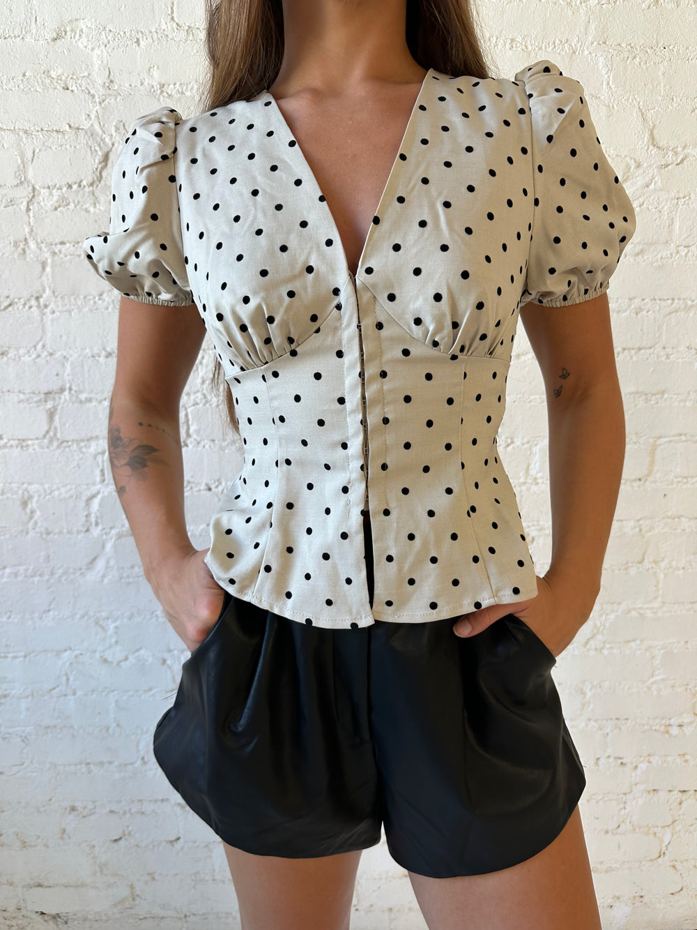 Woman wearing taupe short, puff sleeve, front closure clasp top with black faux leather shorts. 