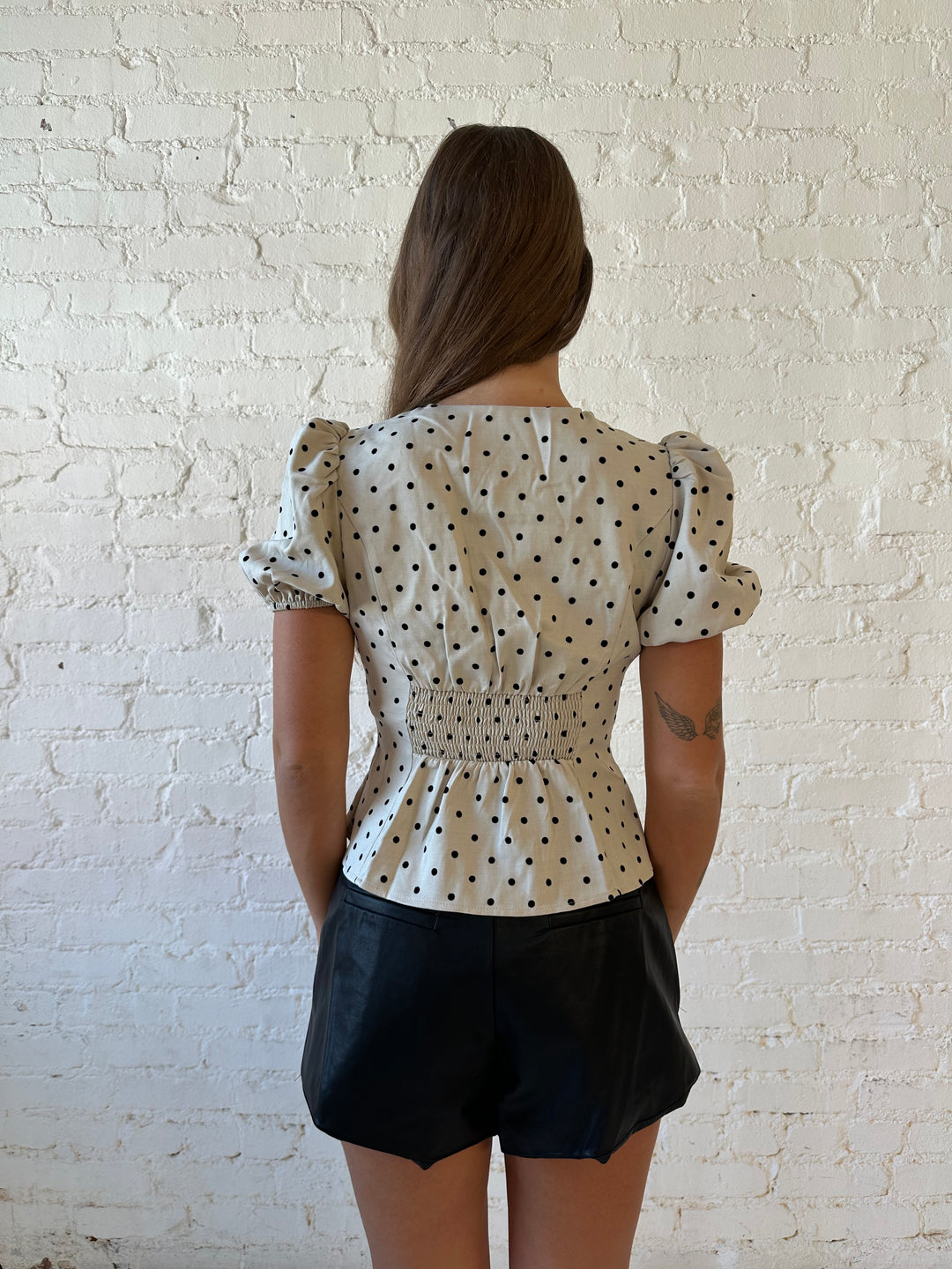 Woman wearing taupe short, puff sleeve, front closure clasp top with black faux leather shorts. 