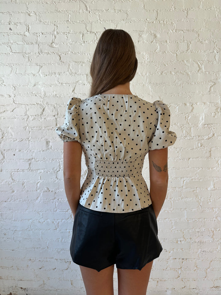 Woman wearing taupe short, puff sleeve, front closure clasp top with black faux leather shorts. 