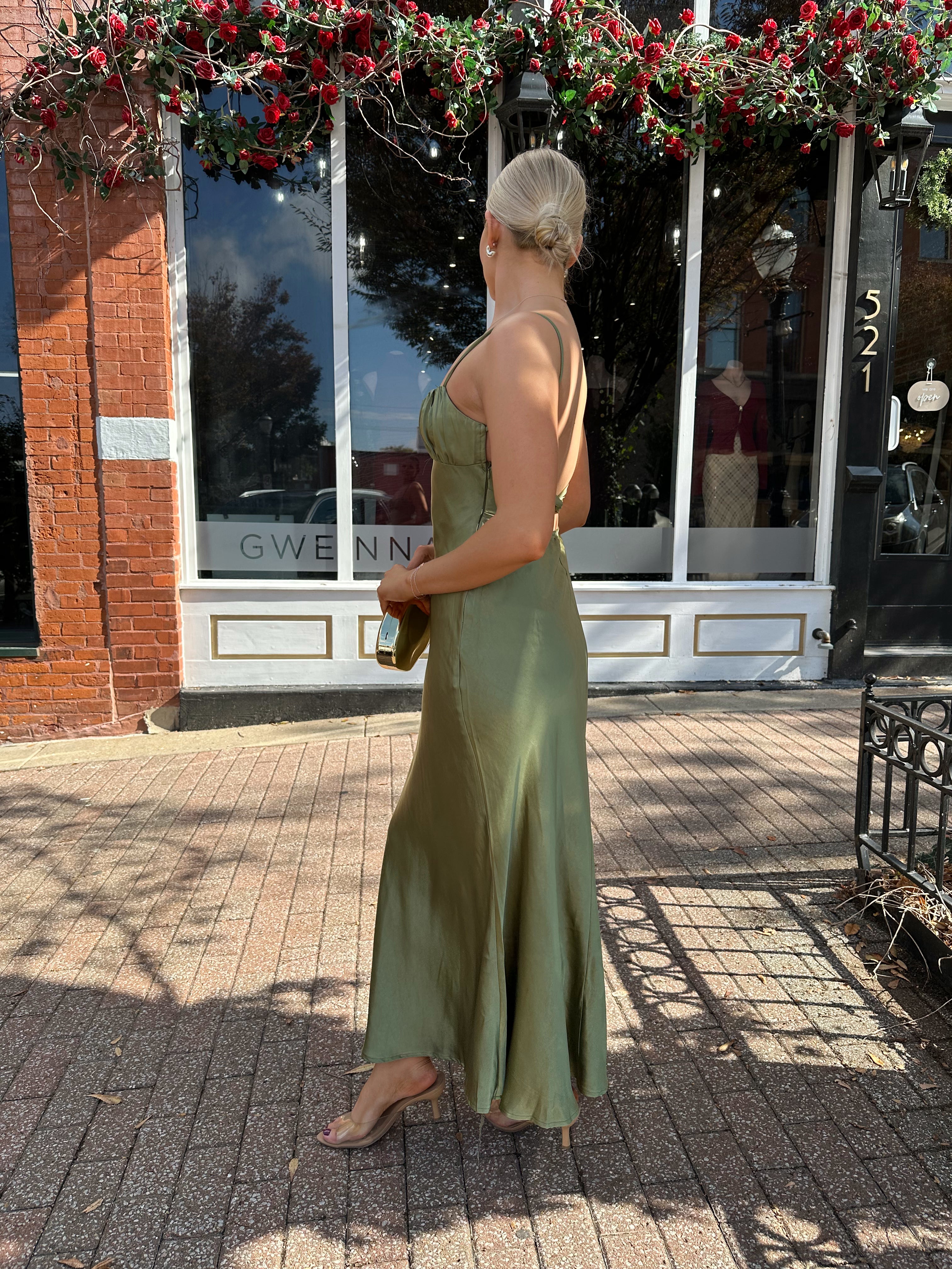 Woman wearing olive green satin maxi dress with open twist back.
