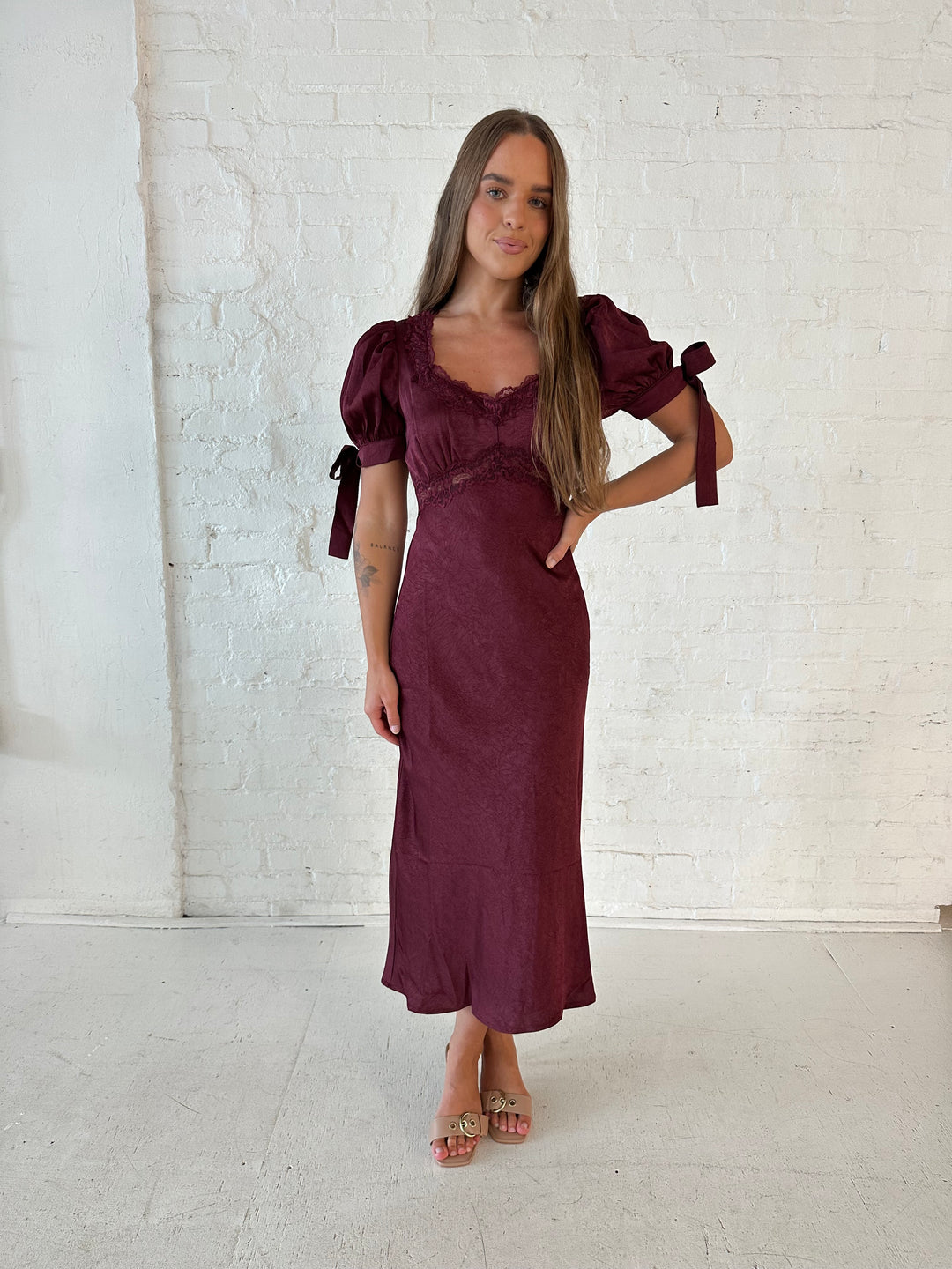 Woman wearing burgundy midi dress with lace trim detailing, and puff sleeves with tie detailing. 