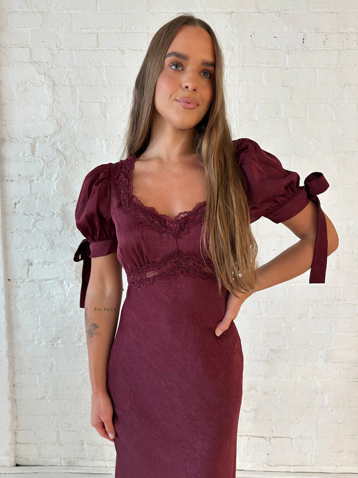 Woman wearing burgundy midi dress with lace trim detailing, and puff sleeves with tie detailing. 