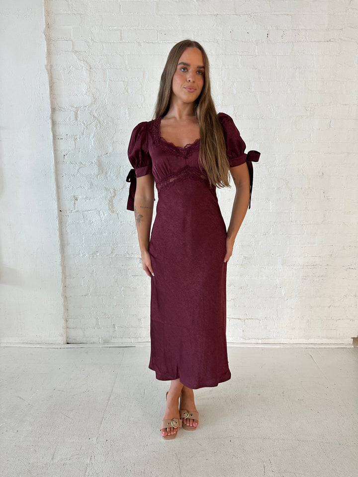 Woman wearing burgundy midi dress with lace trim detailing, and puff sleeves with tie detailing. 