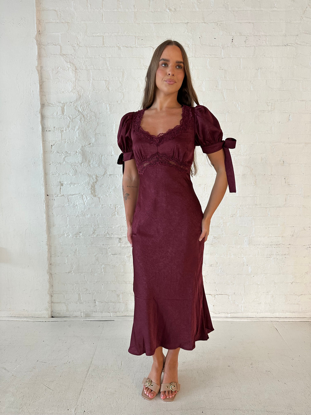 Woman wearing burgundy midi dress with lace trim detailing, and puff sleeves with tie detailing. 
