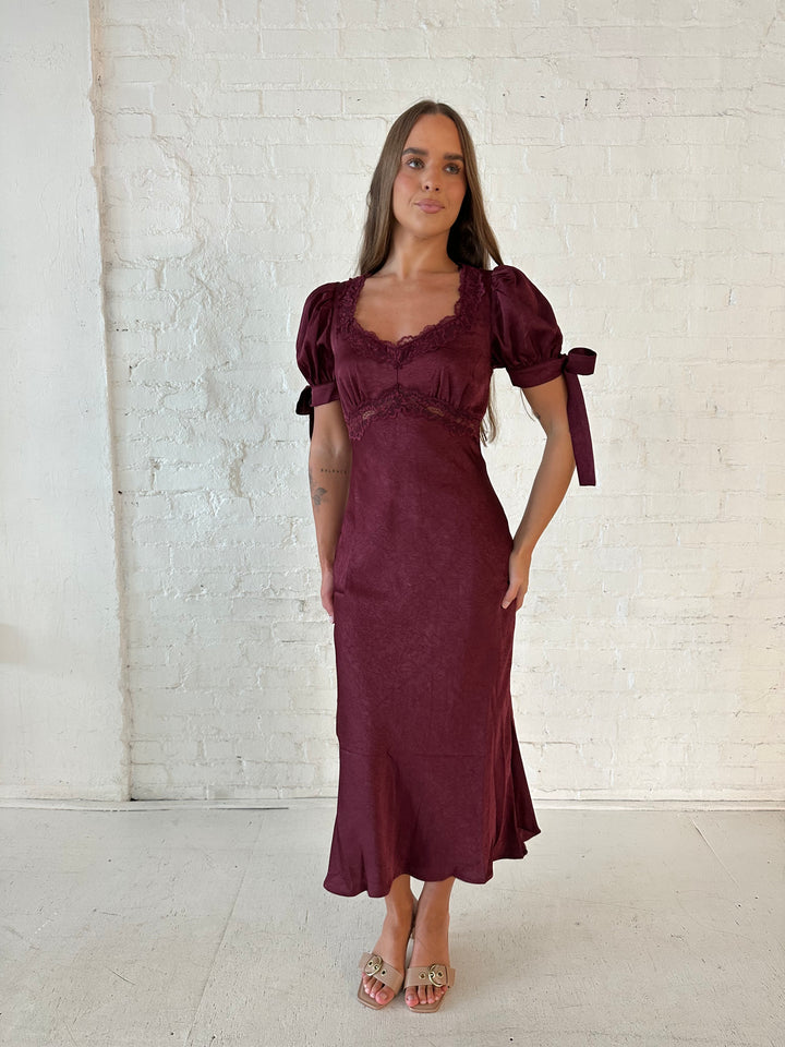 Woman wearing burgundy midi dress with lace trim detailing, and puff sleeves with tie detailing. 