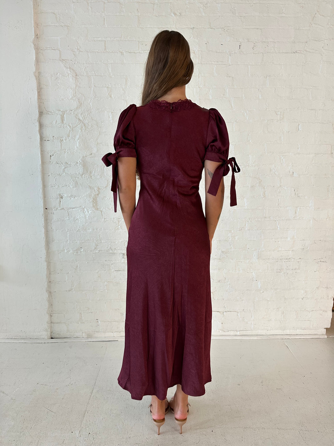 Woman wearing burgundy midi dress with lace trim detailing, and puff sleeves with tie detailing. 