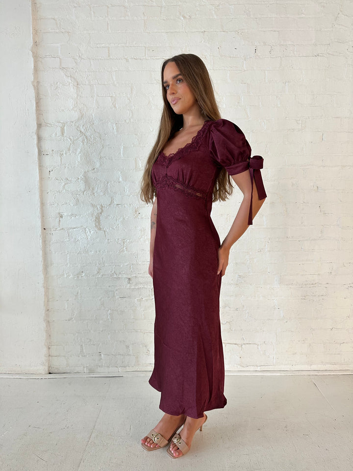 Woman wearing burgundy midi dress with lace trim detailing, and puff sleeves with tie detailing. 