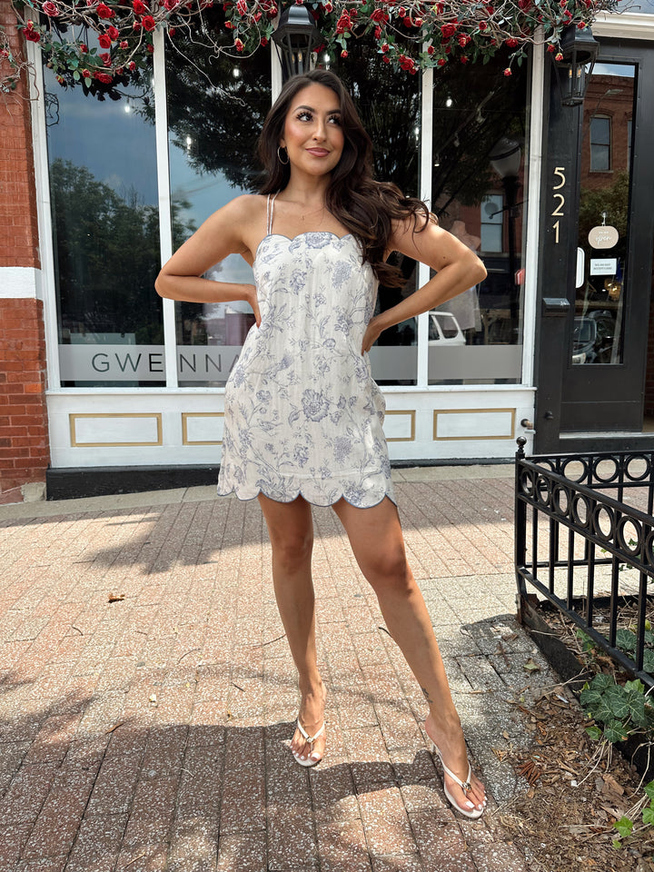 Woman wearing off white mini dress with blue floral pattern, scallop hem, and adjustable tie straps