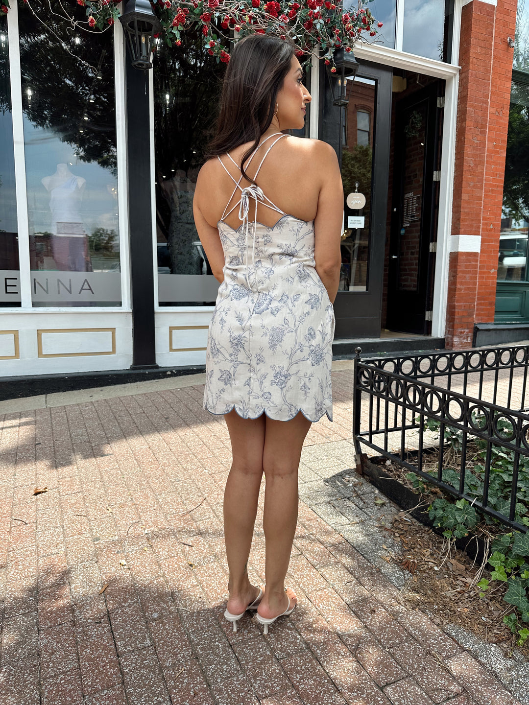 Woman wearing off white mini dress with blue floral pattern, scallop hem, and adjustable tie straps