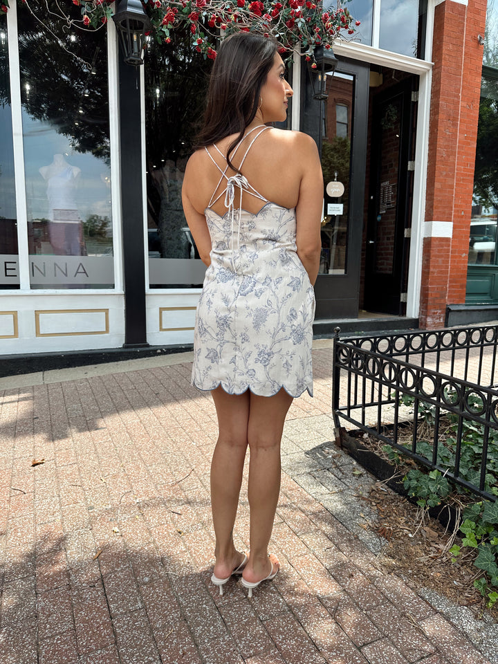 Woman wearing off white mini dress with blue floral pattern, scallop hem, and adjustable tie straps