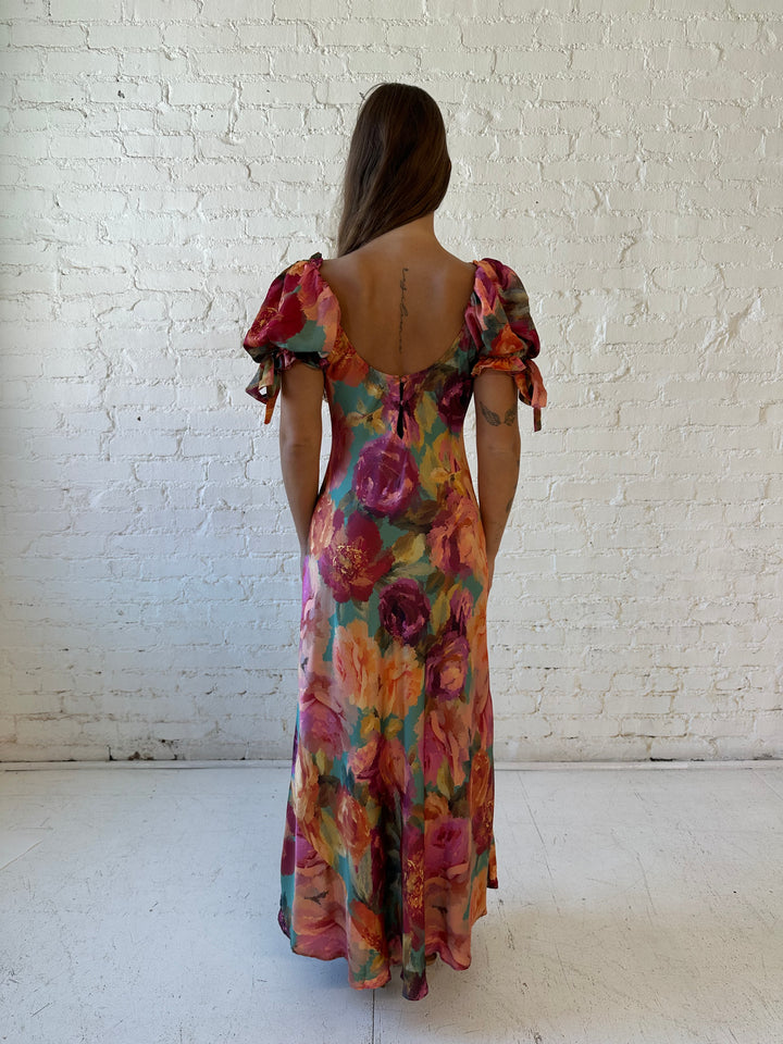 Woman wearing multicolor floral satin maxi dress with puff sleeves and tie detail.