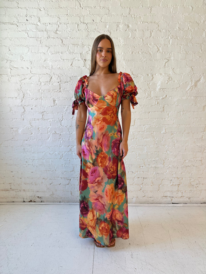 Woman wearing multicolor floral satin maxi dress with puff sleeves and tie detail.