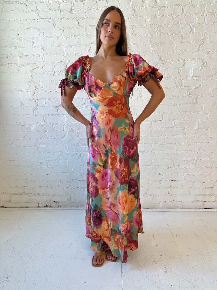 Woman wearing multicolor floral satin maxi dress with puff sleeves and tie detail.