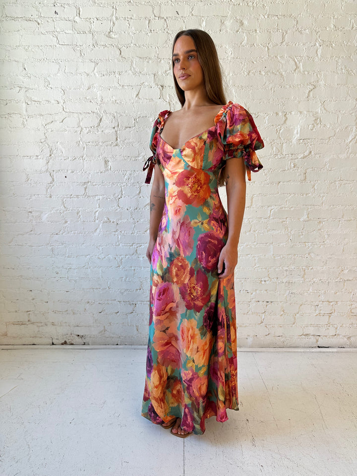Woman wearing multicolor floral satin maxi dress with puff sleeves and tie detail.