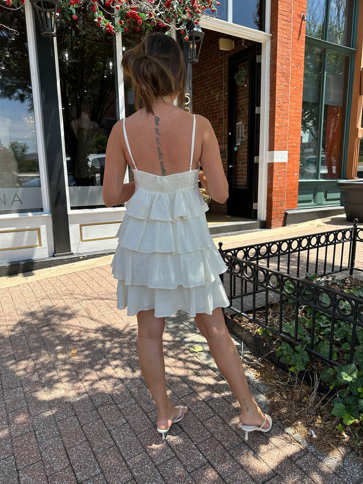 Woman wearing beige tiered mini dress with v-neckline, and beaded strap detailing. Has adjustable spaghetti straps.