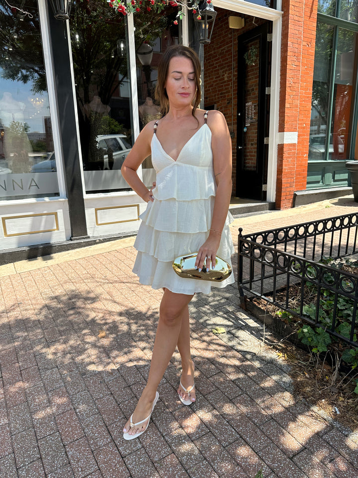 Woman wearing beige tiered mini dress with v-neckline, and beaded strap detailing. Has adjustable spaghetti straps.