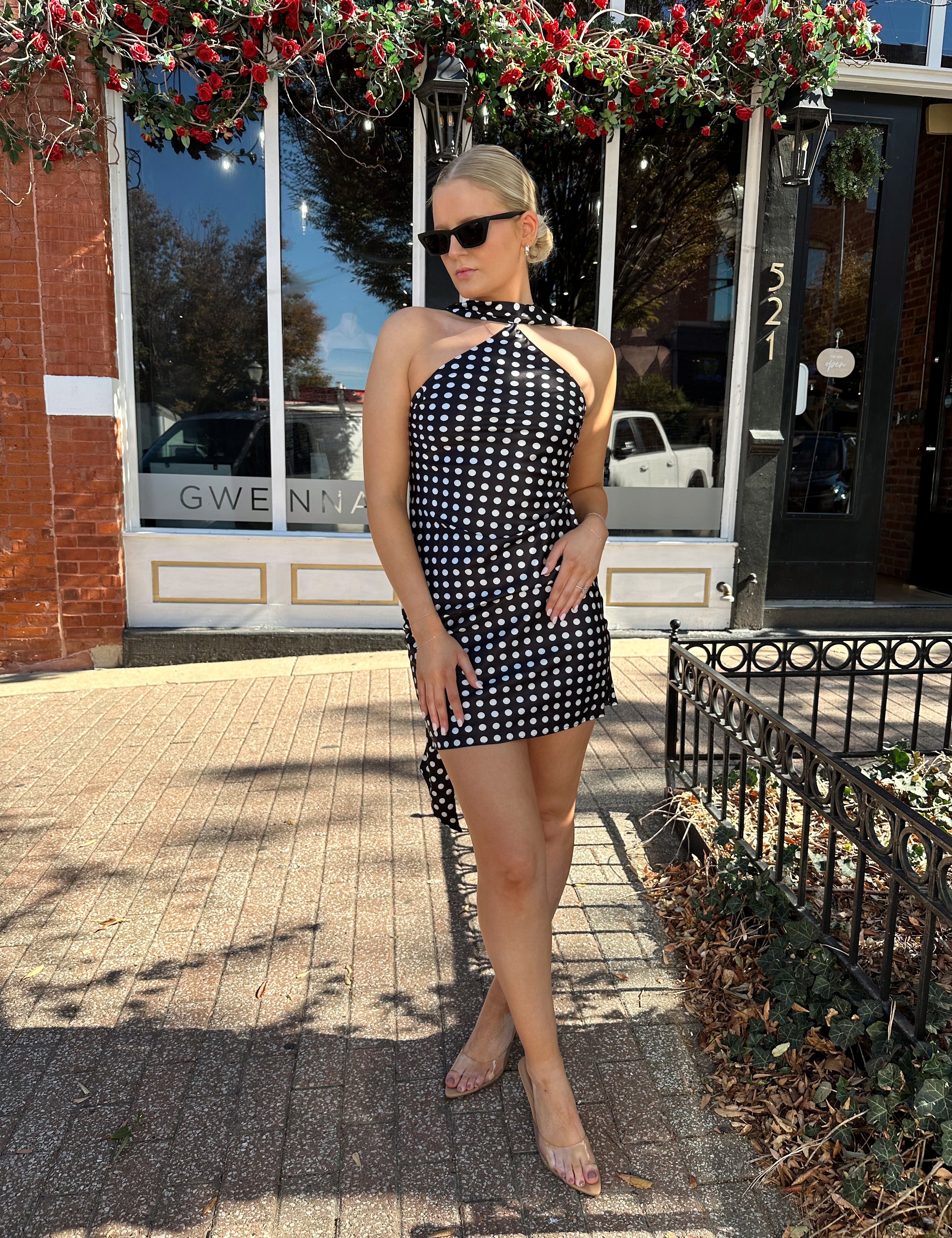 Woman wearing black and white polka dot satin mini dress with halter scarf neckline. 