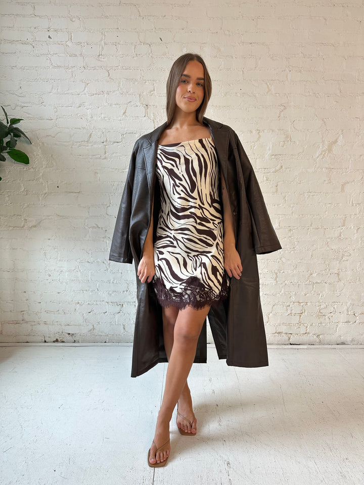 Woman wearing dark brown faux leather trench coat over brown abstract satin mini dress. 