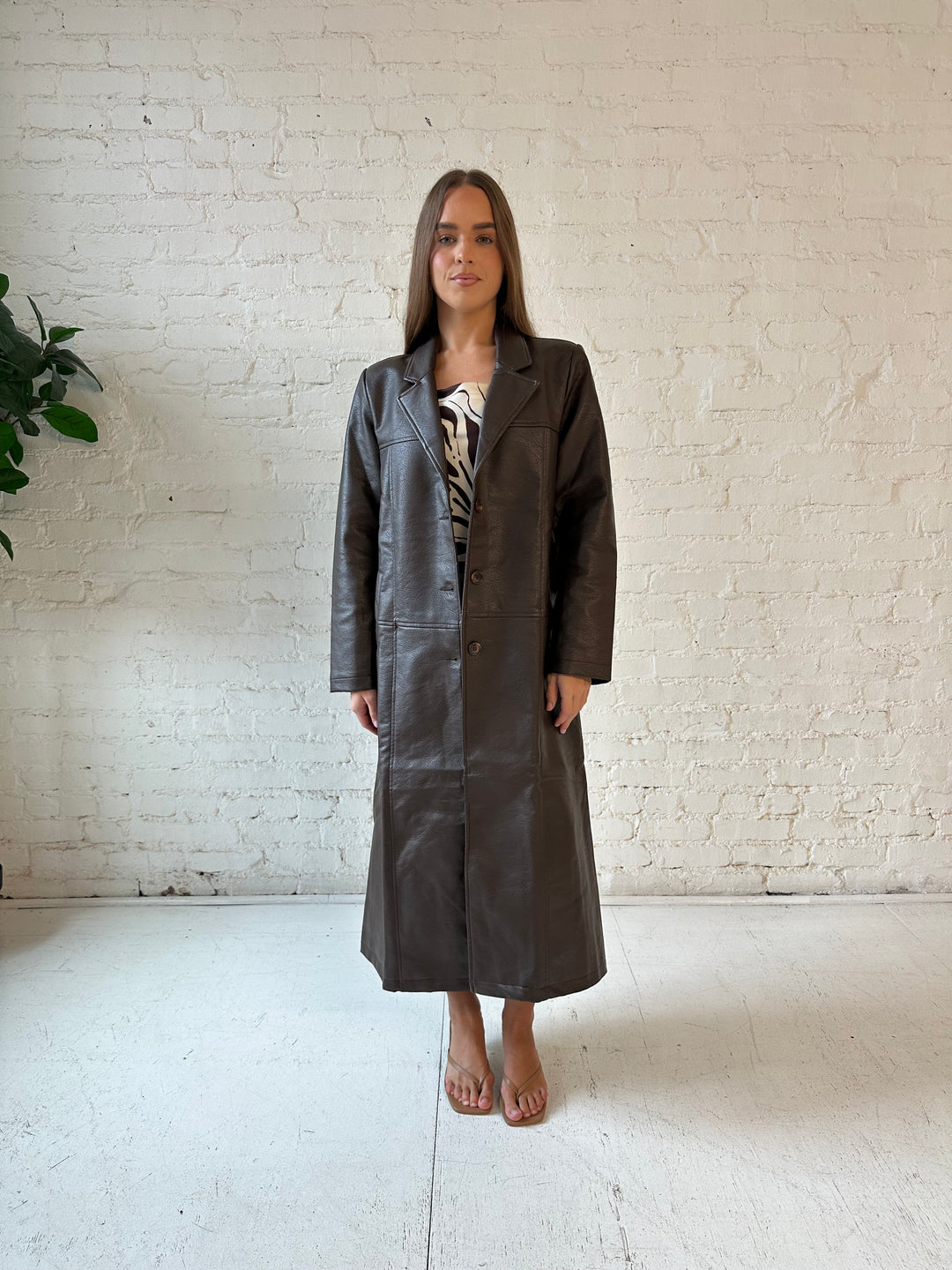 Woman wearing dark brown faux leather trench coat over brown abstract satin mini dress. 