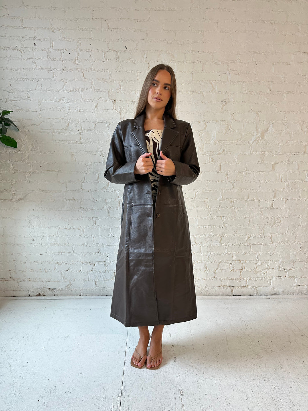 Woman wearing dark brown faux leather trench coat over brown abstract satin mini dress. 