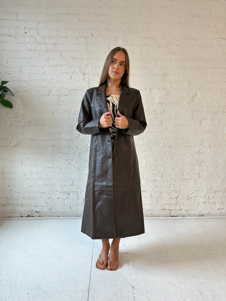 Woman wearing dark brown faux leather trench coat over brown abstract satin mini dress. 