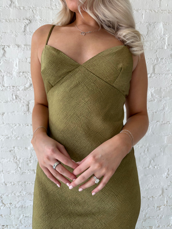 Woman wearing olive green maxi dress with v-neckline and adjustable straps. 