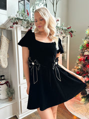 Woman wearing black velvet babydoll mini dress with puff sleeves, and bow detailing. 