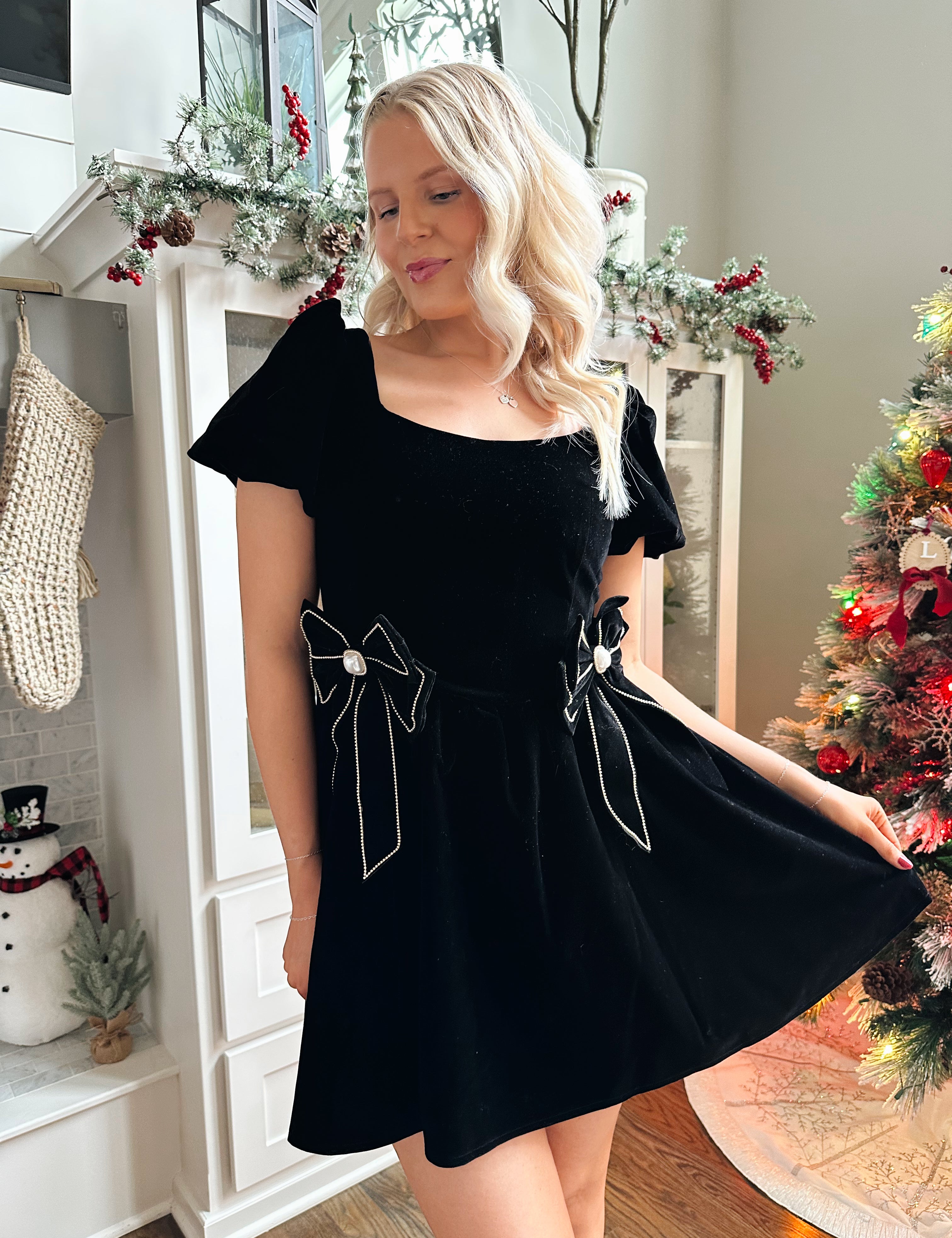 Woman wearing black velvet babydoll mini dress with puff sleeves, and bow detailing. 