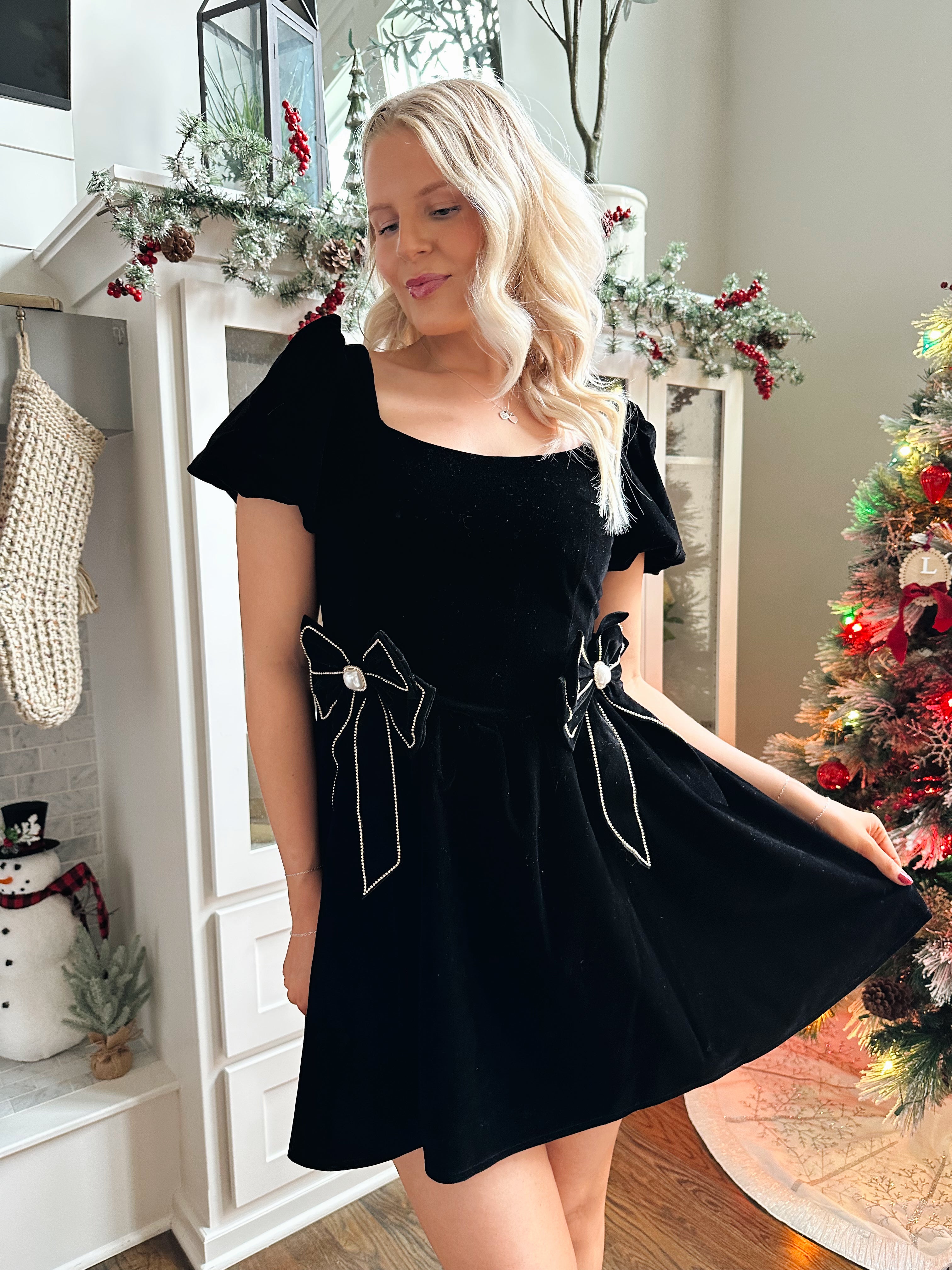Woman wearing black velvet babydoll mini dress with puff sleeves, and bow detailing. 