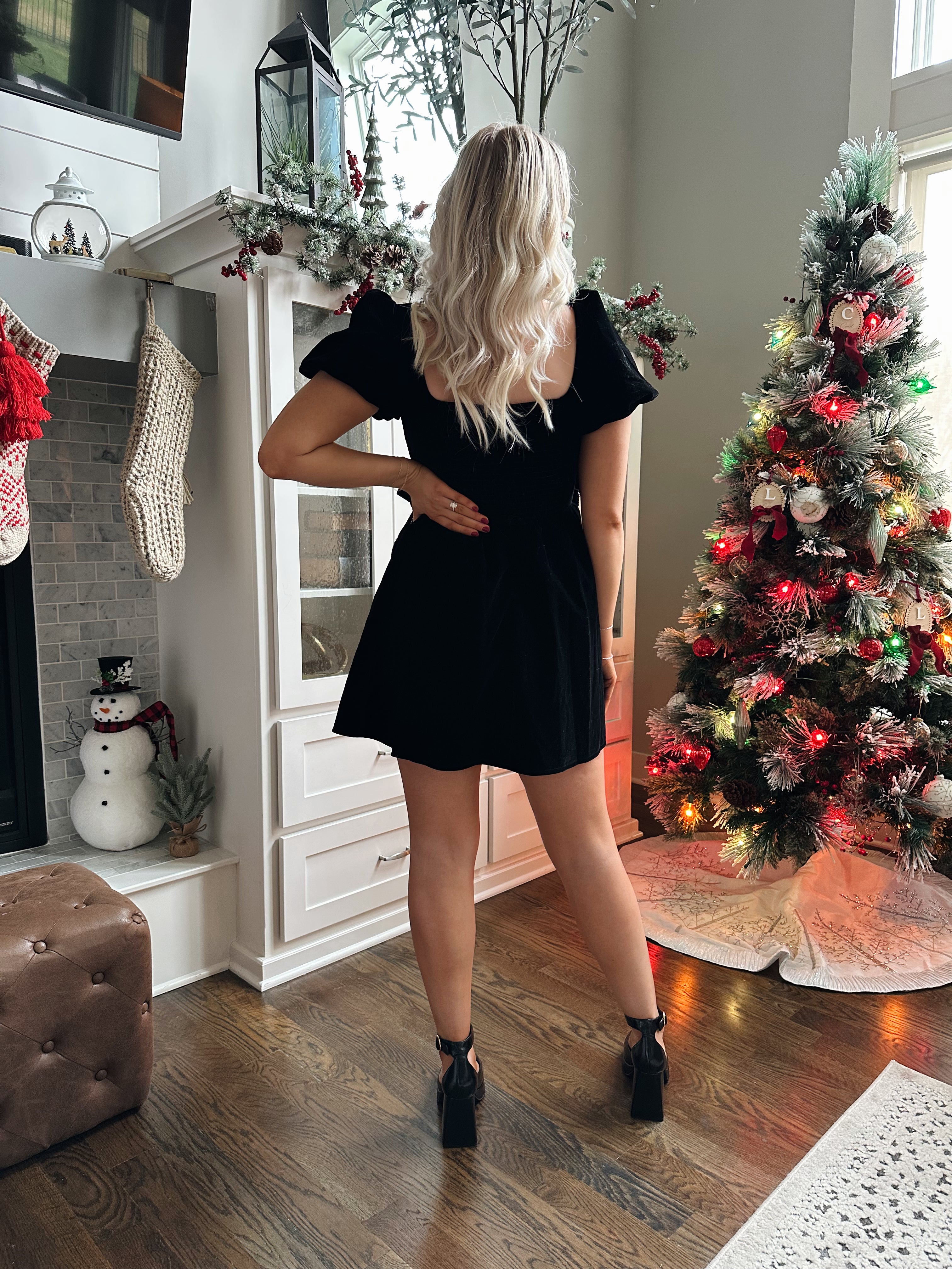 Woman wearing black velvet babydoll mini dress with puff sleeves, and bow detailing. 