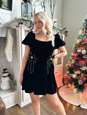 Woman wearing black velvet babydoll mini dress with puff sleeves, and bow detailing. 