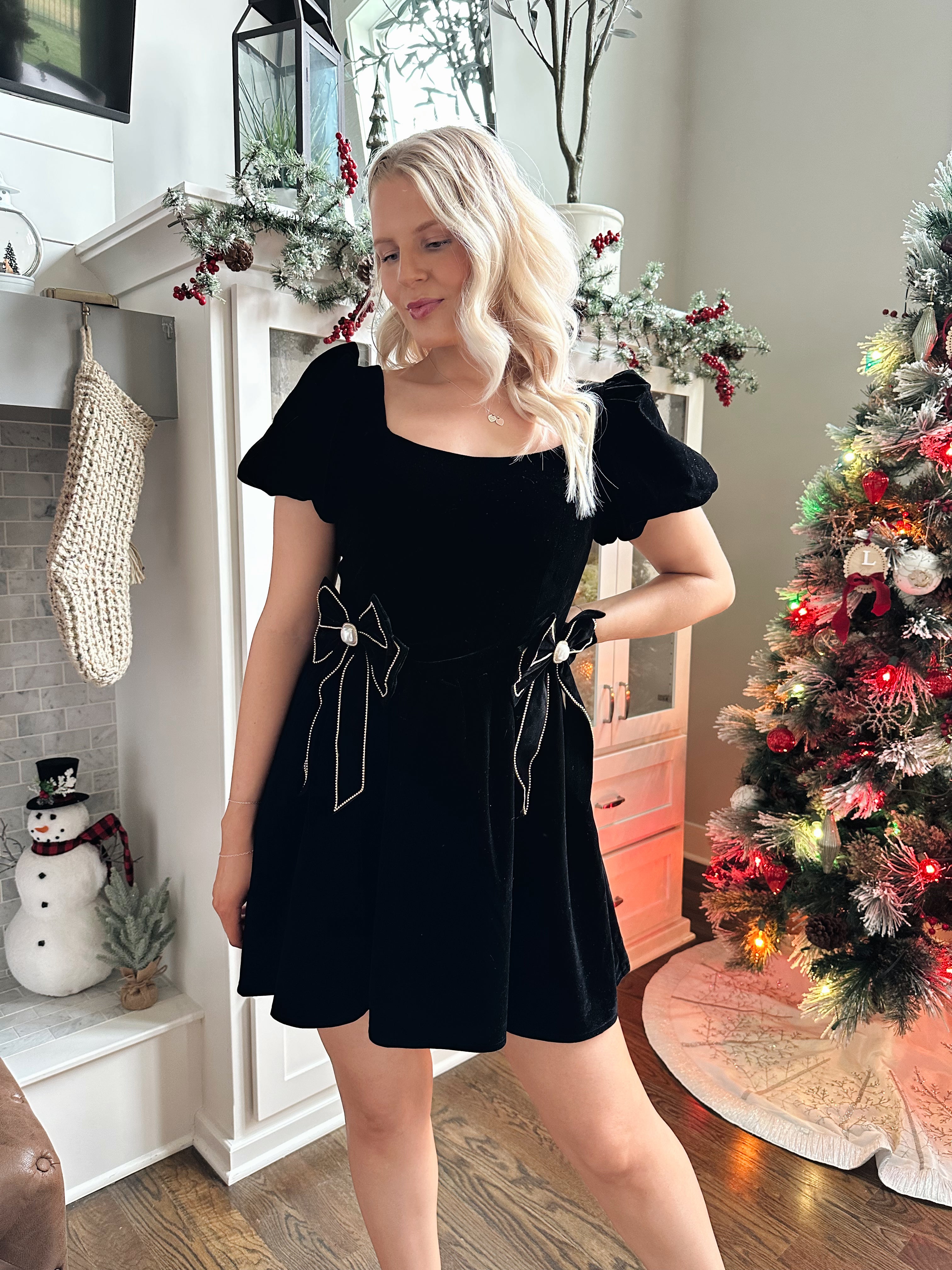 Woman wearing black velvet babydoll mini dress with puff sleeves, and bow detailing. 