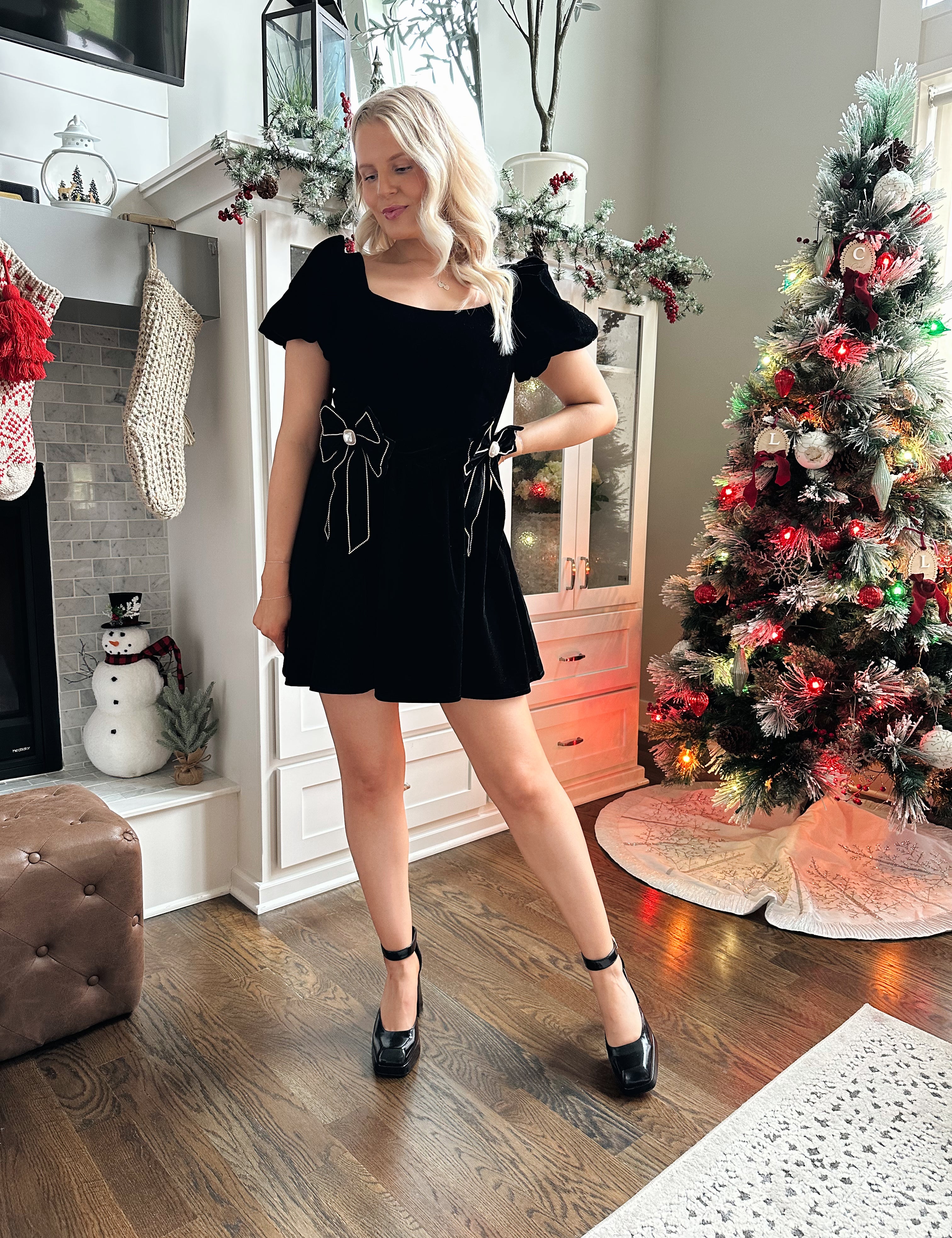 Woman wearing black velvet babydoll mini dress with puff sleeves, and bow detailing. 