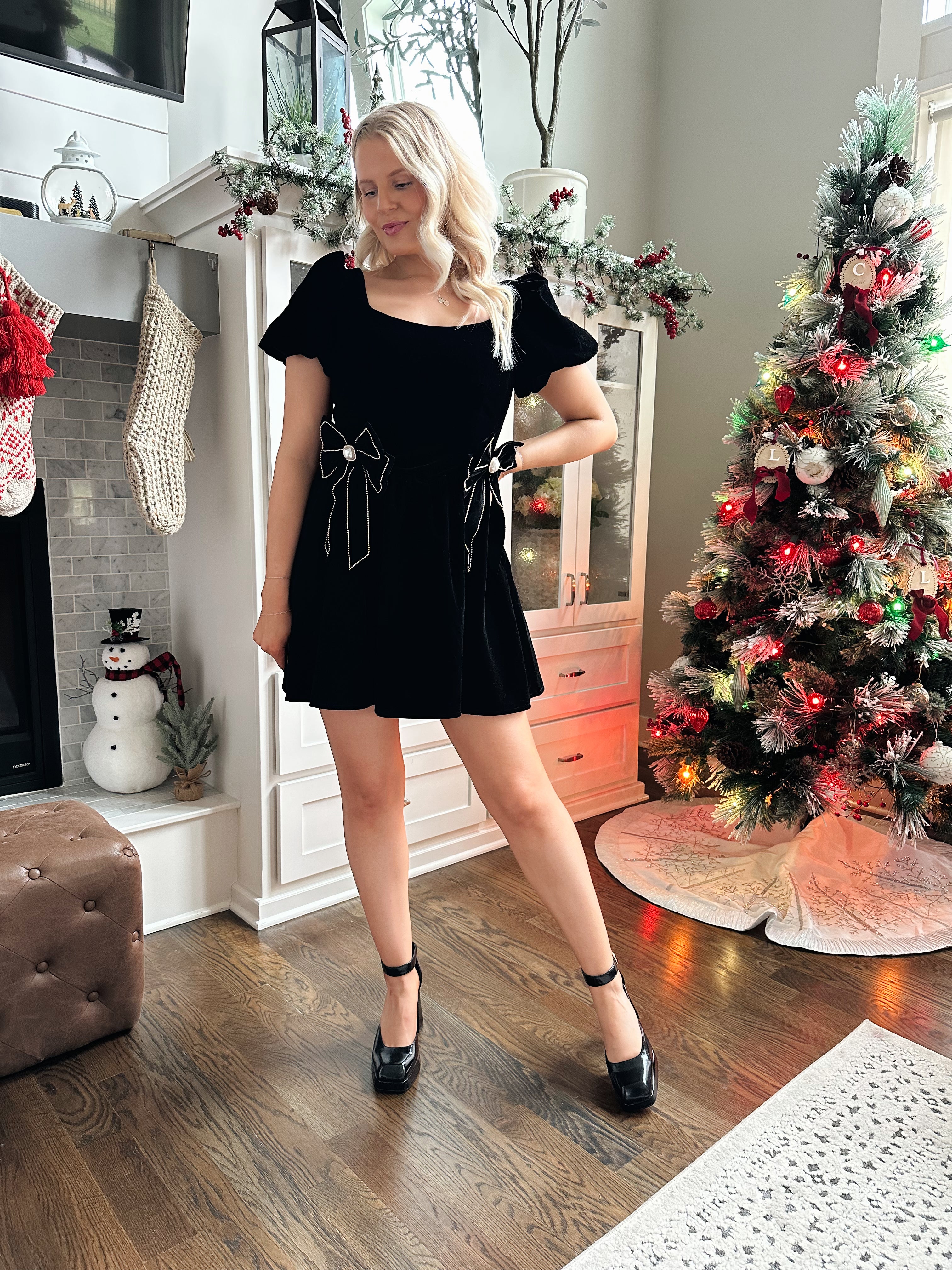 Woman wearing black velvet babydoll mini dress with puff sleeves, and bow detailing. 