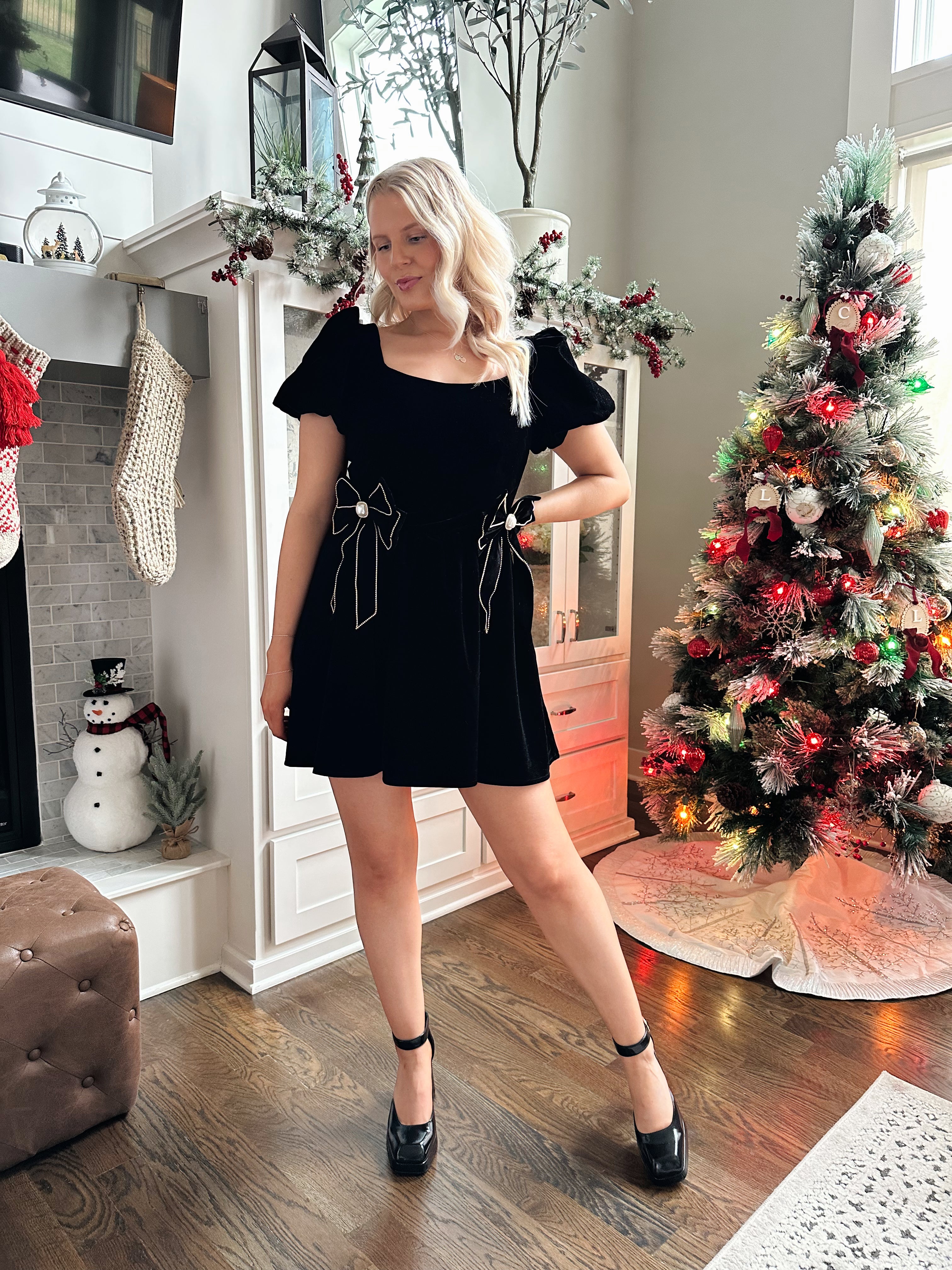 Woman wearing black velvet babydoll mini dress with puff sleeves, and bow detailing. 